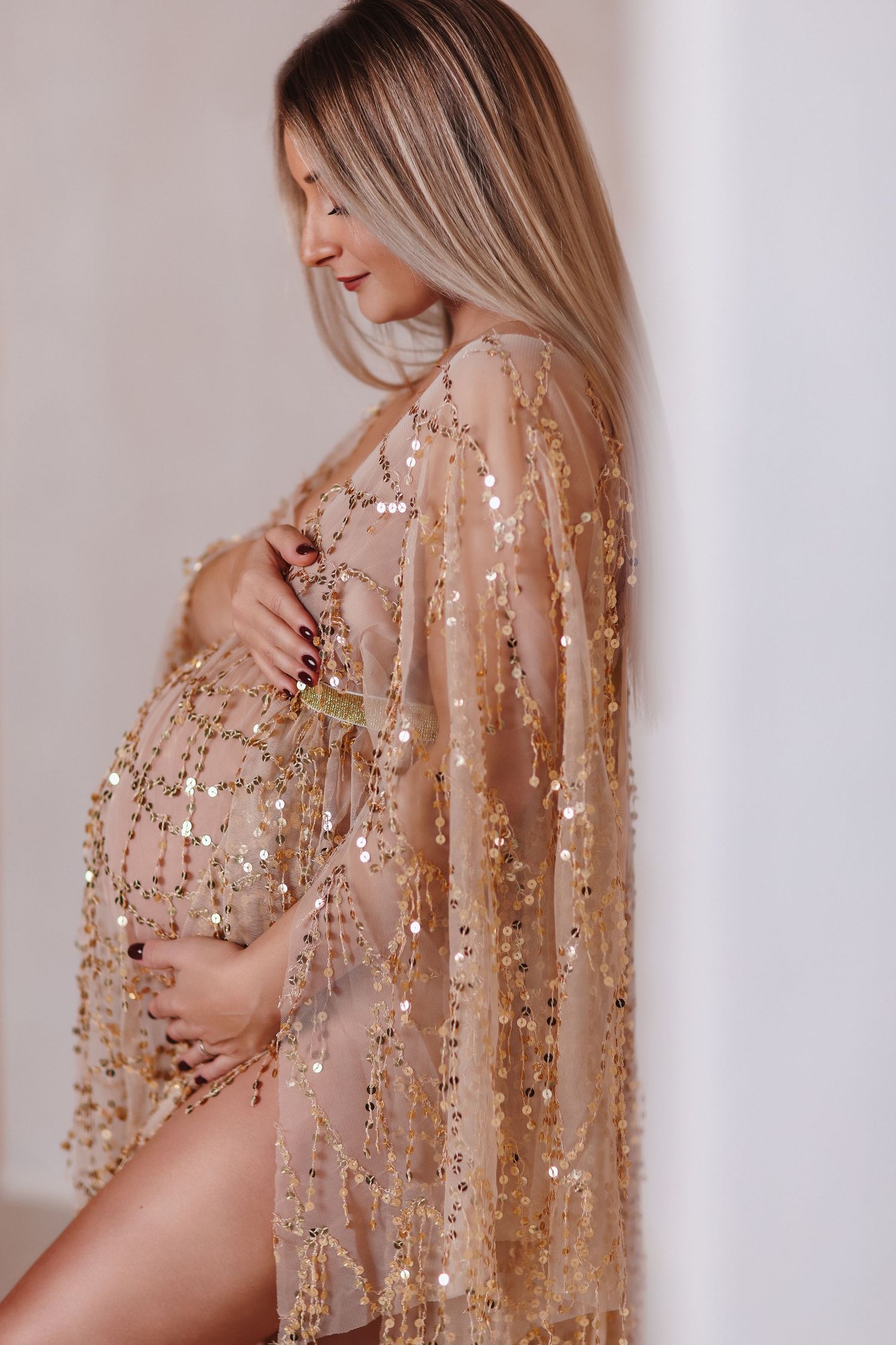 Pregnant woman in gold sequin sheer robe cradling baby bump, elegant maternity photography studio portrait.
