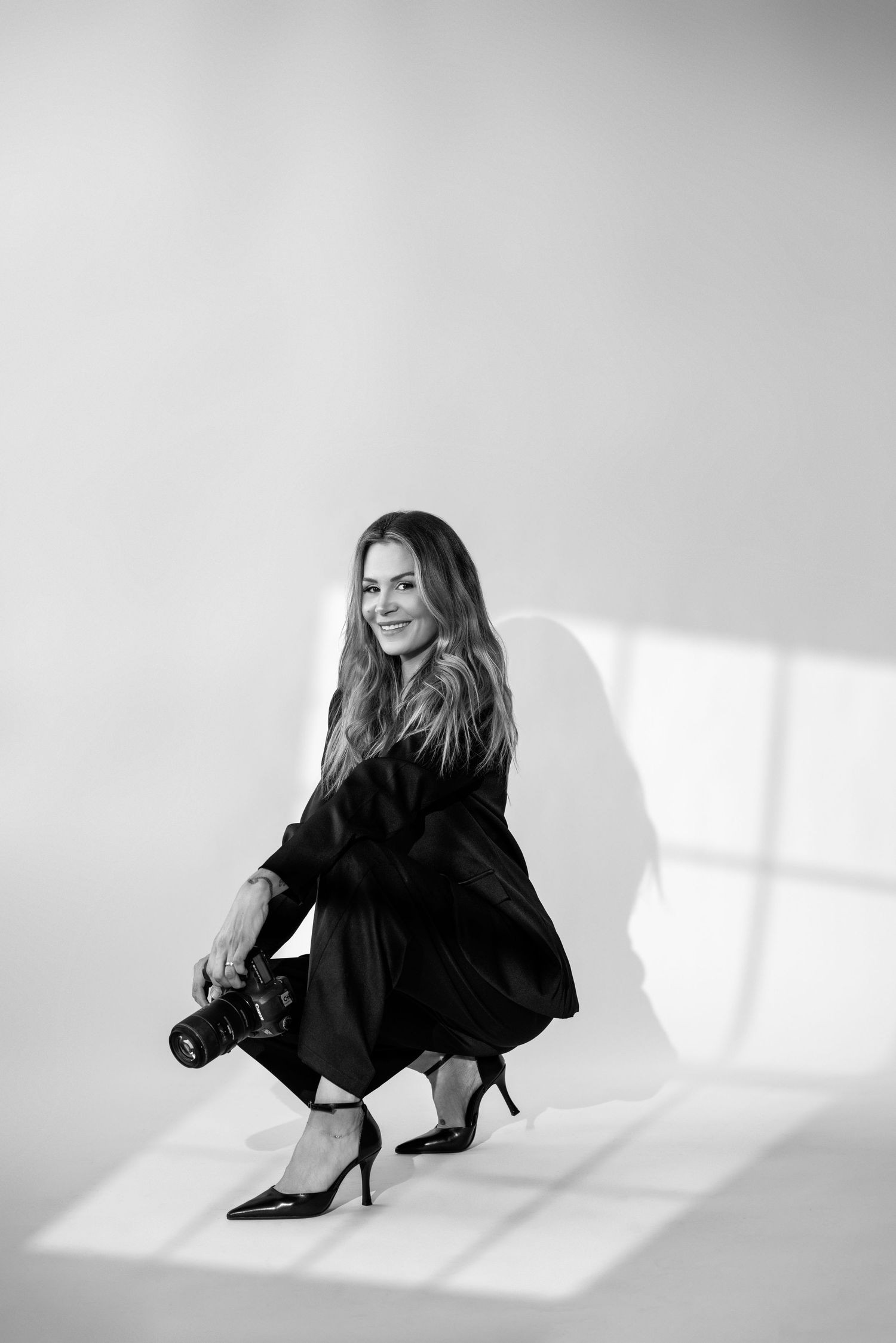 Black and white photo of a smiling woman in a black dress holding a camera, crouching in a bright studio.