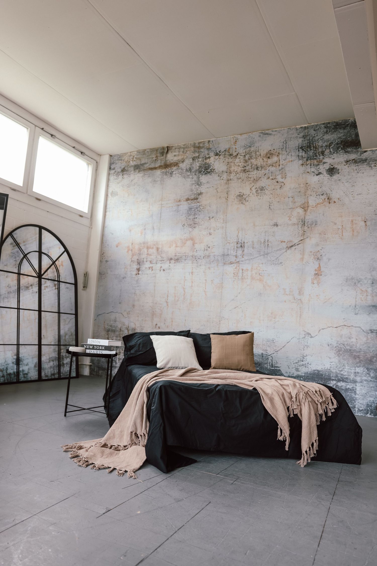 Industrial bedroom with black bedding, beige throw, and distressed wall art in a loft-style space with concrete floors.