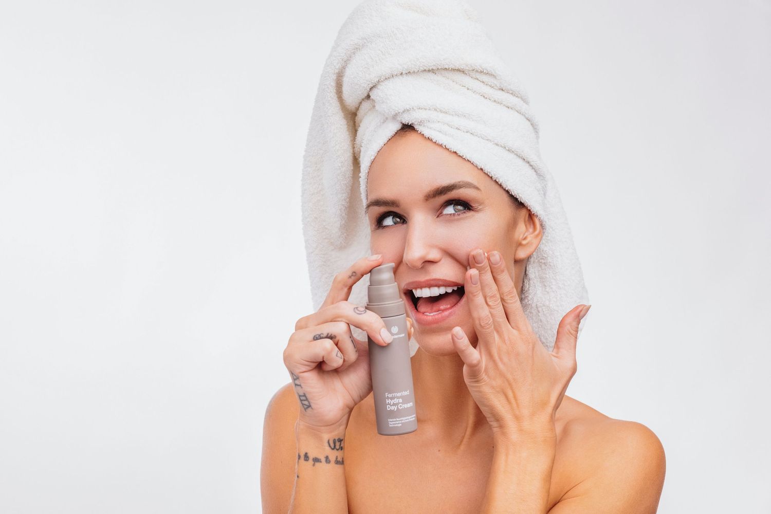 Woman with towel on head applying facial serum, smiling against white background.