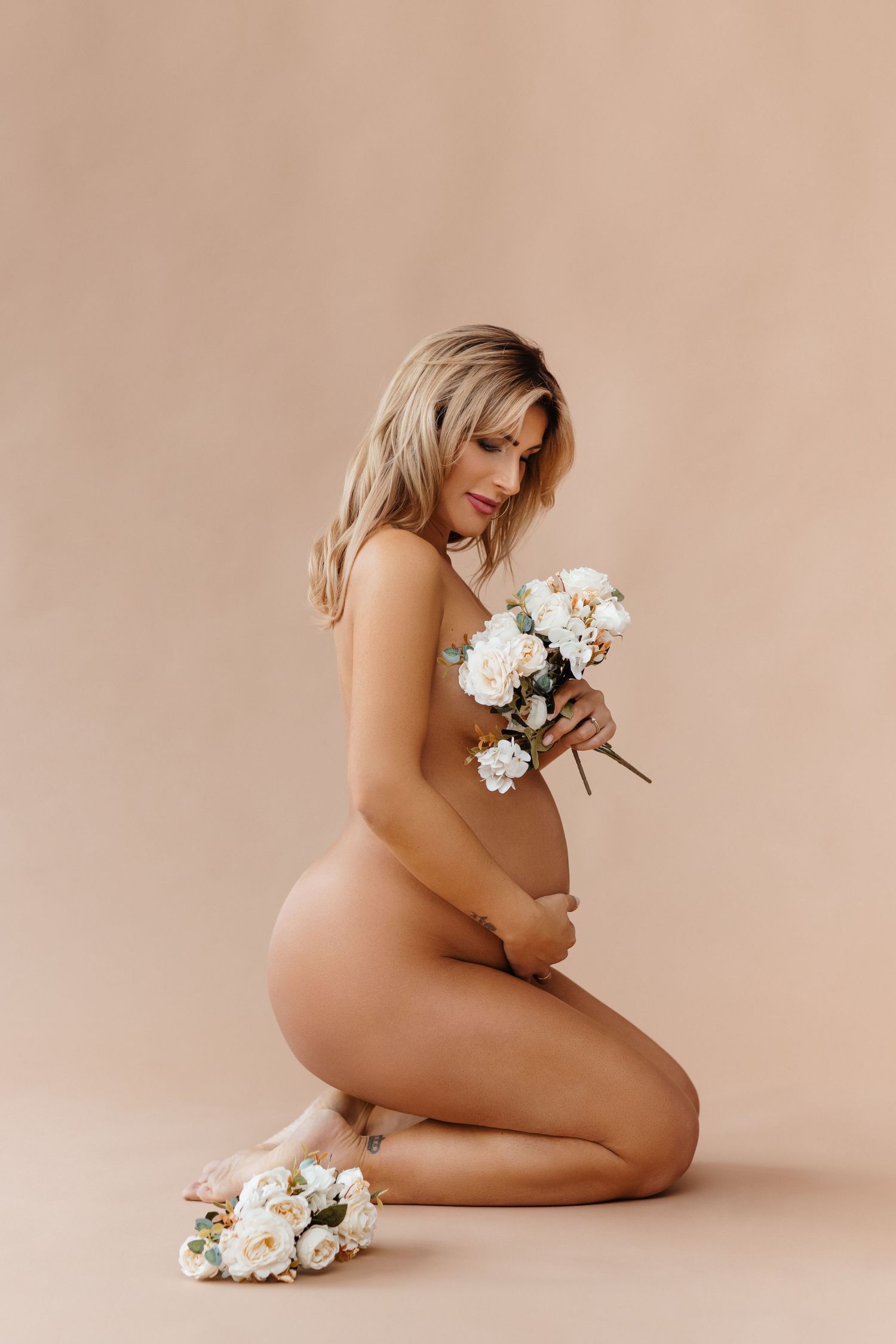 Pregnant woman kneeling nude holding white flowers against beige background in elegant maternity portrait.