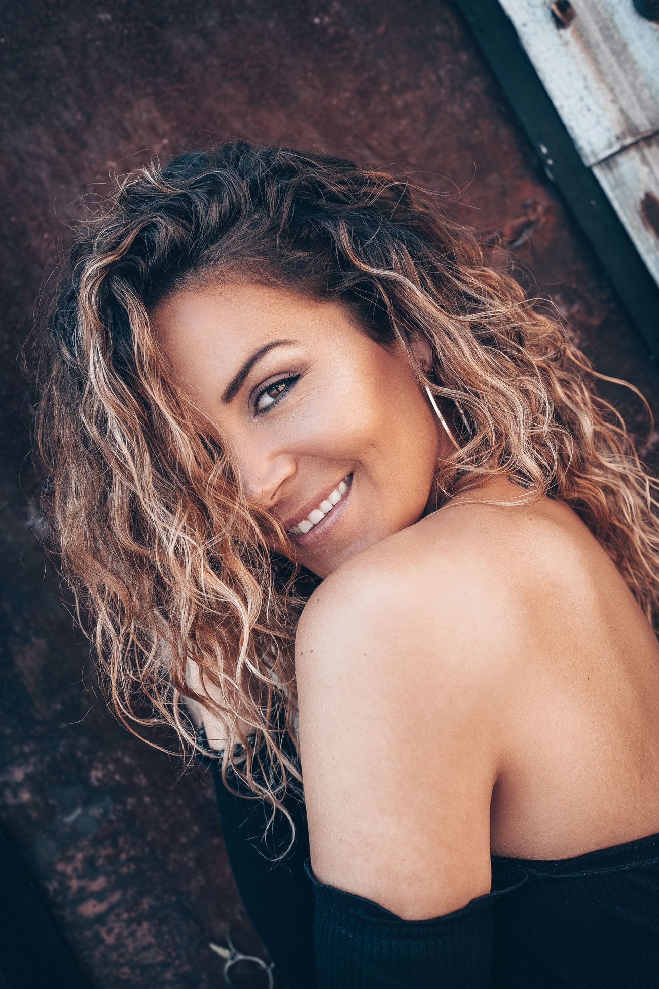 Smiling woman with curly blonde highlighted hair wearing a black top, posing against a rustic metal background.