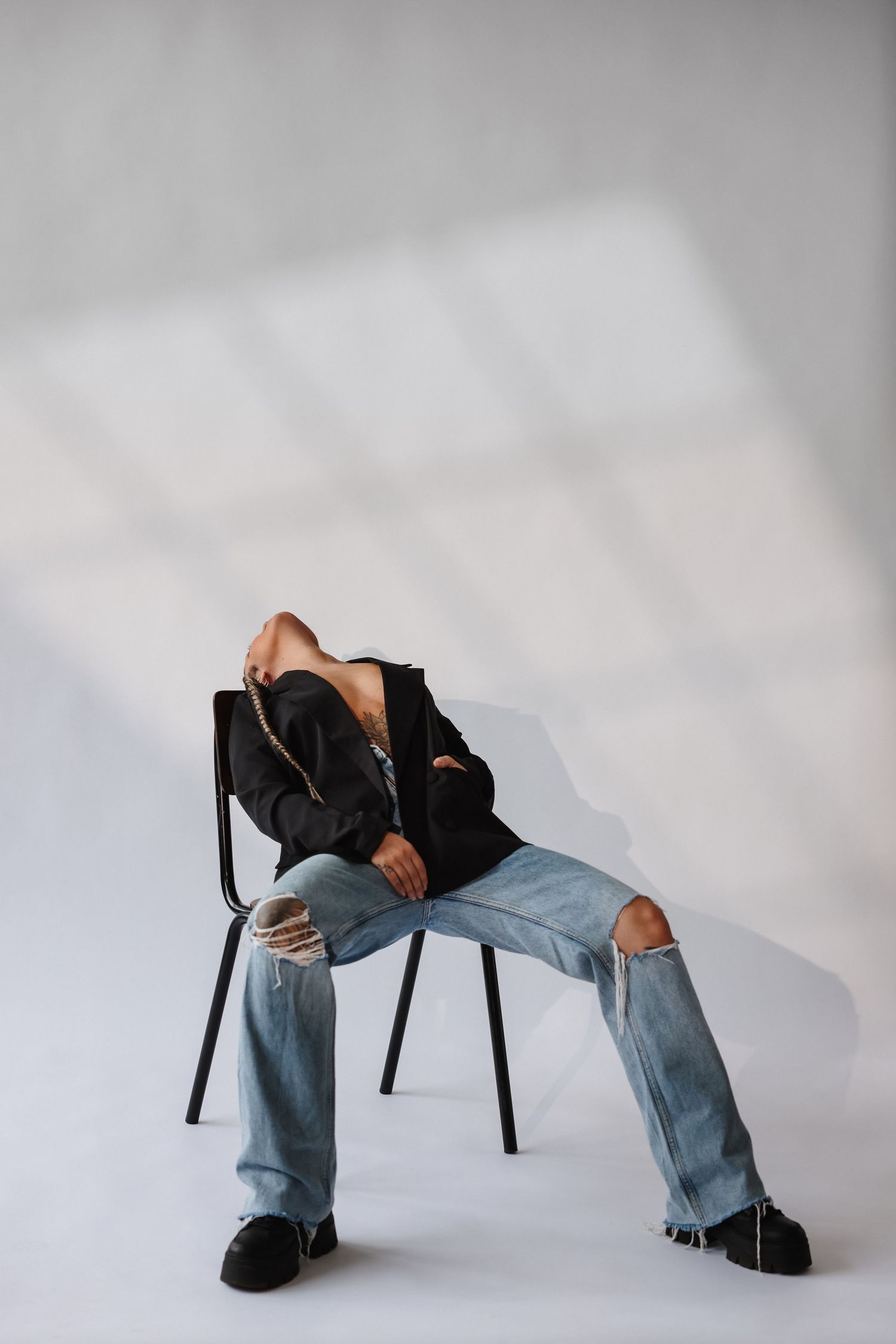Person sitting on black chair in ripped jeans and black blazer, head tilted back, against white wall with light shadows.