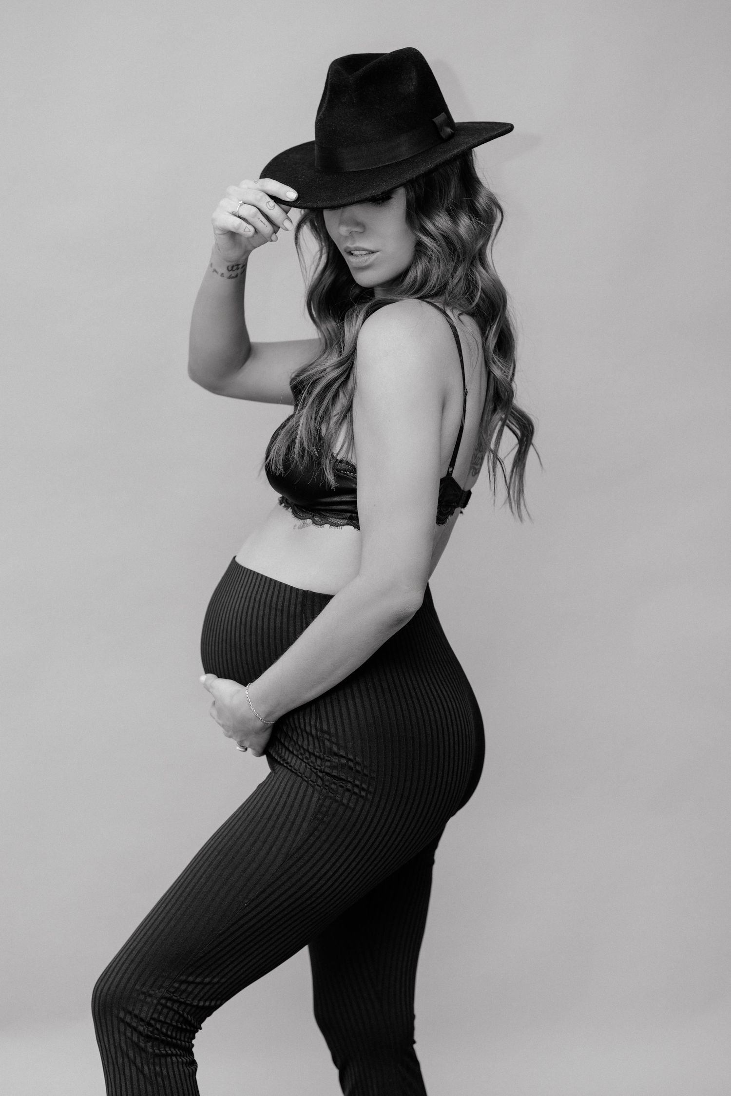 Black and white maternity photo of a woman in a crop top, striped pants, and wide-brim hat cradling her baby bump.