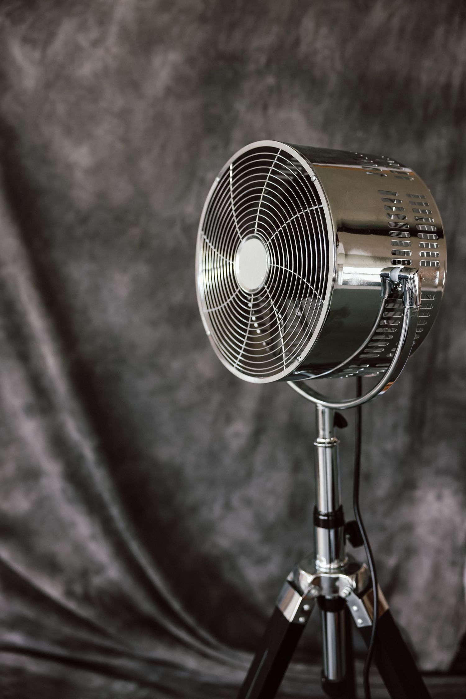 A retro chrome electric fan on a tripod stand against a dark gray mottled studio backdrop.