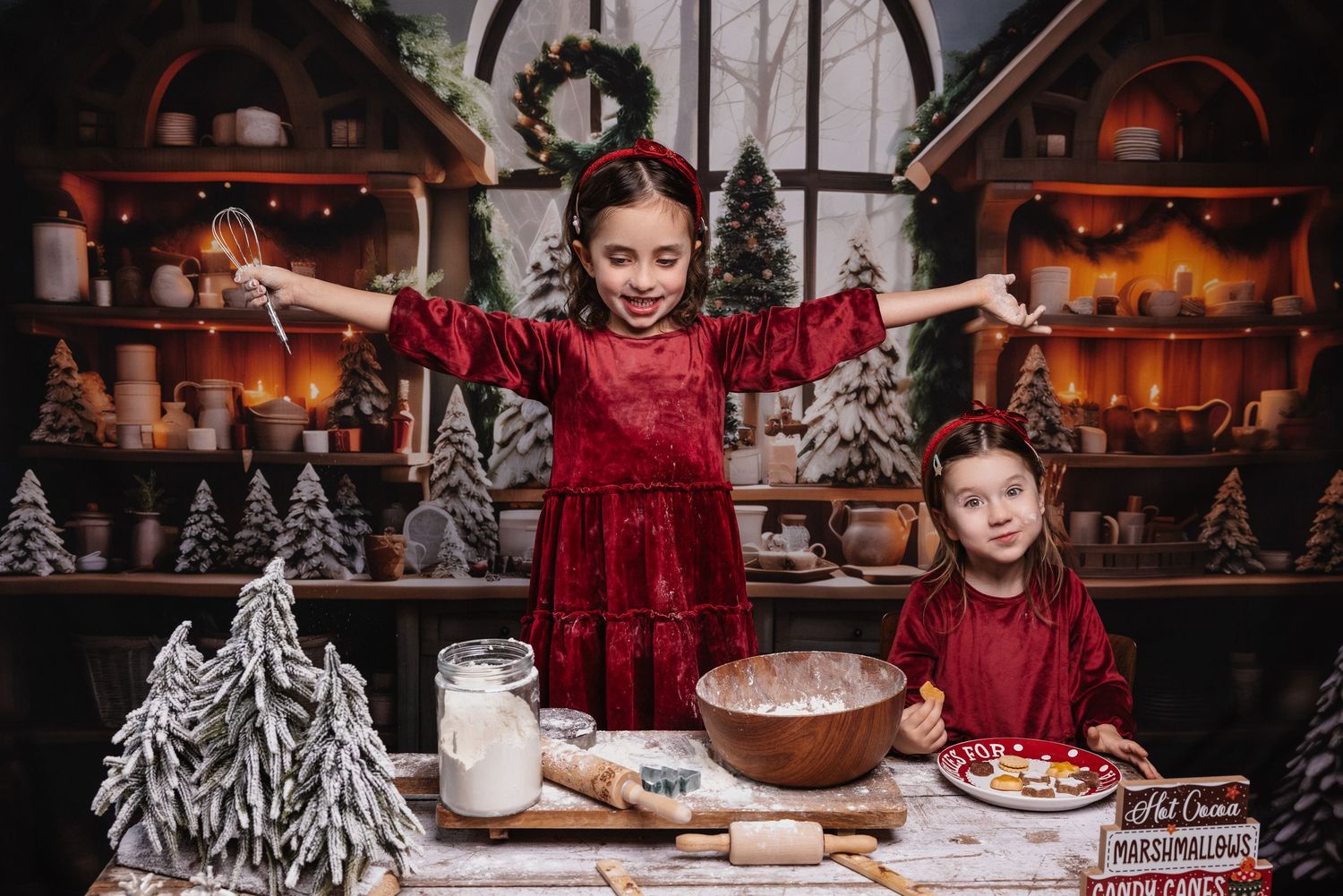 Two girls in red velvet dresses bake Christmas cookies at a festive decorated table with holiday trees and shelves.