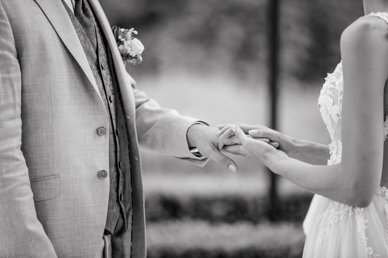 Black and white photo of groom placing wedding ring on bride's finger during outdoor ceremony.