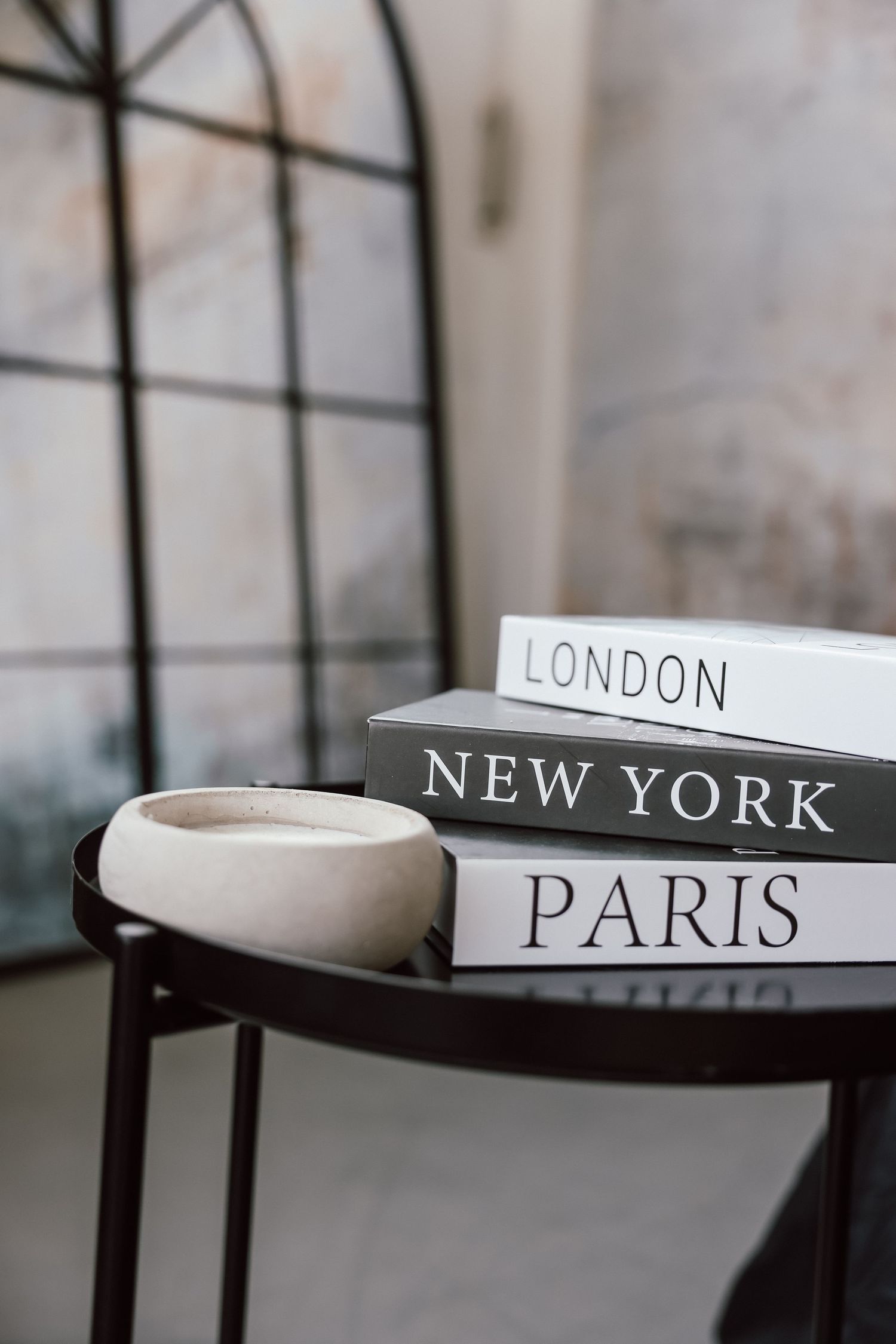 Stack of London, New York, and Paris books on a dark side table with a ceramic bowl and arched mirror backdrop.