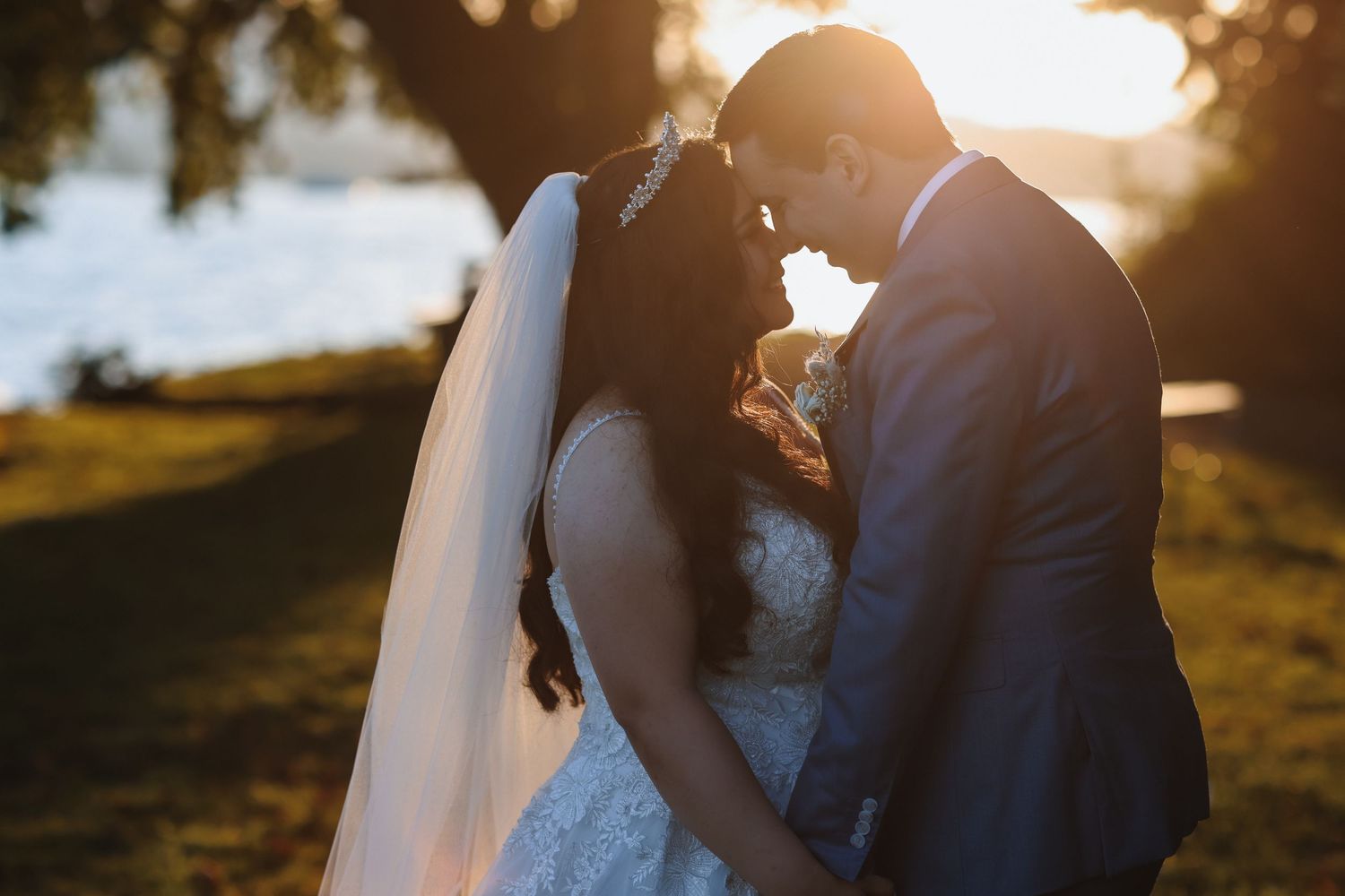 Bride and groom sharing a romantic forehead touch at sunset, with golden light glowing behind them near a waterfront.