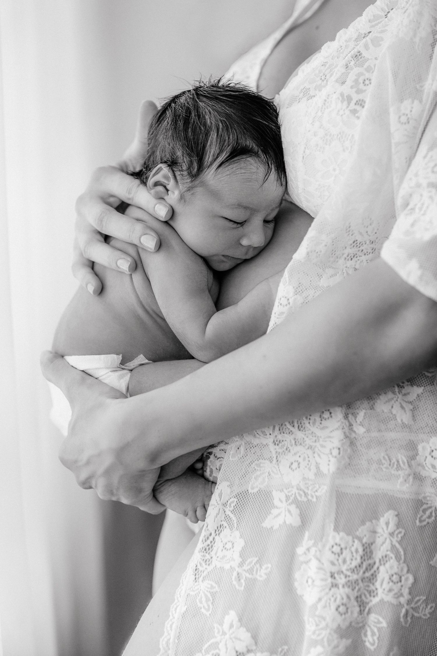 Black and white photo of a mother in lace dress cradling her sleeping newborn baby close to her chest.