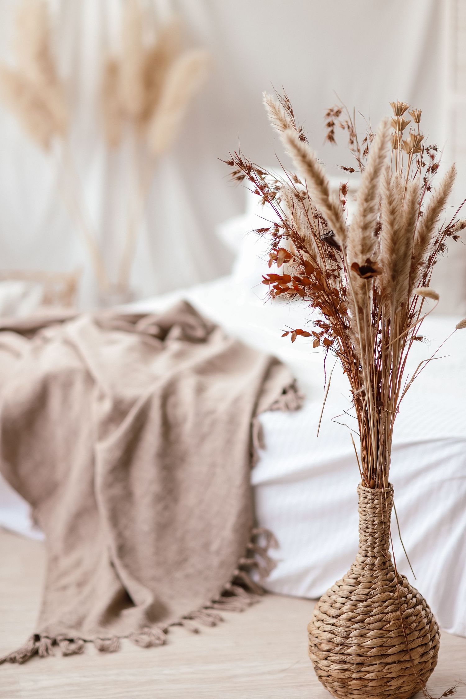 Woven vase with dried pampas grass and wildflowers on a bed with beige linen throw in a boho bedroom.