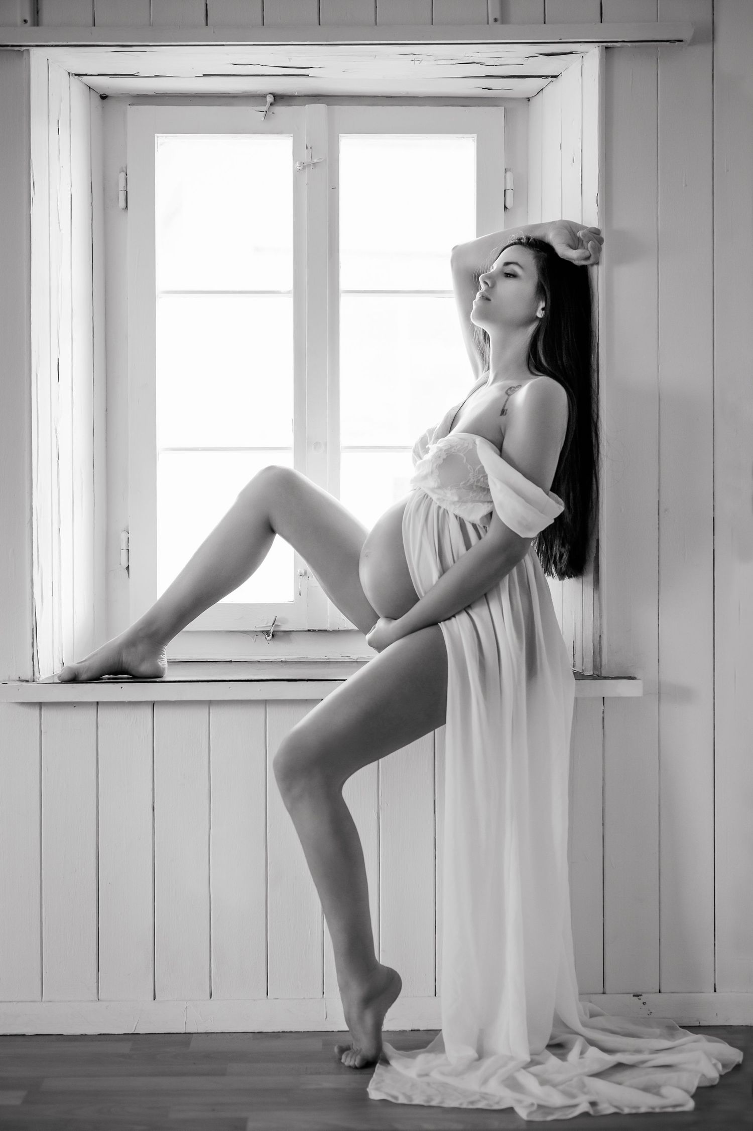 Black and white photo of a pregnant woman in white sheer dress posing gracefully on a windowsill.