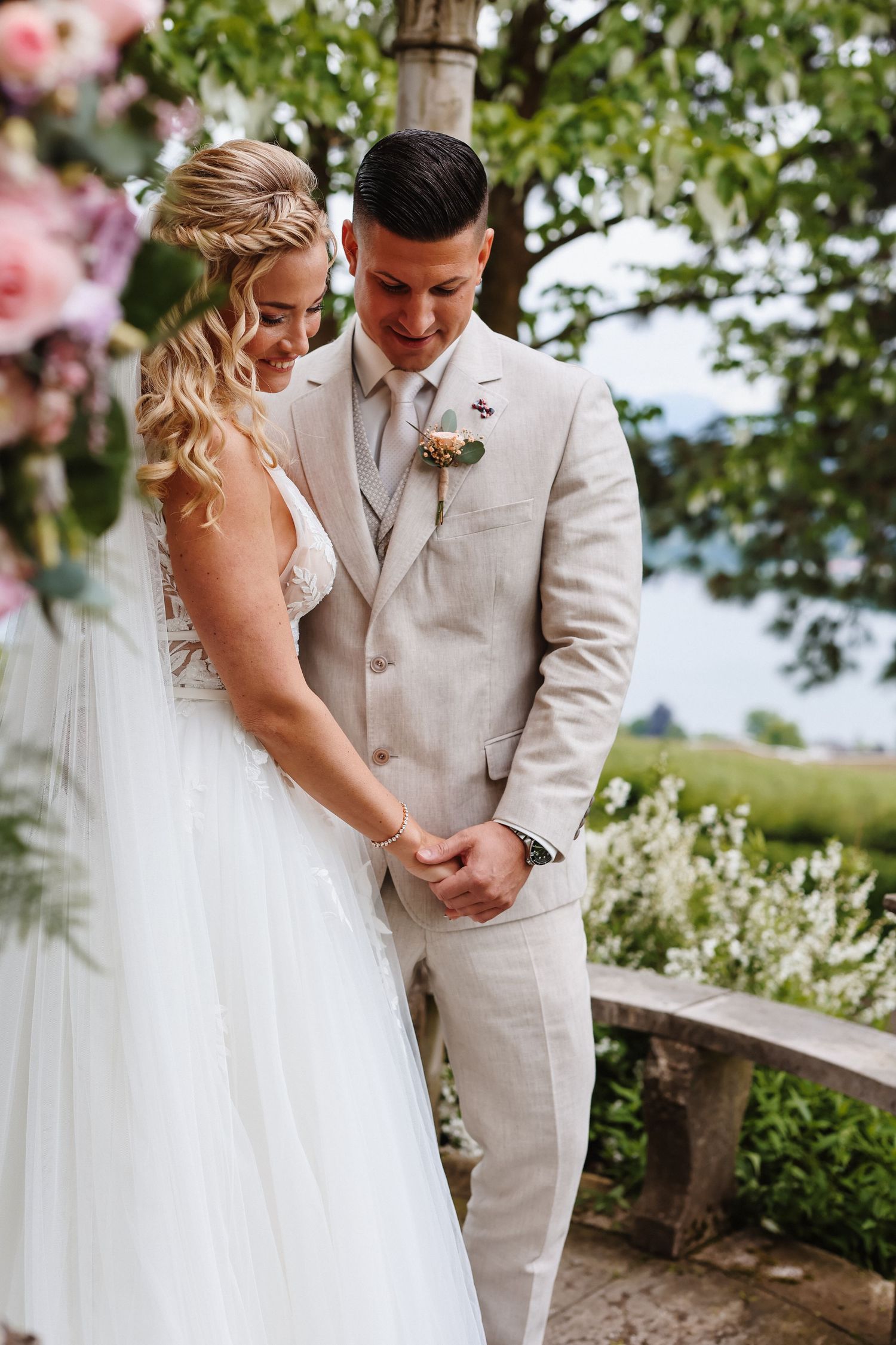 Bride and groom holding hands outdoors at wedding ceremony, groom in beige suit, bride in white gown, floral backdrop.