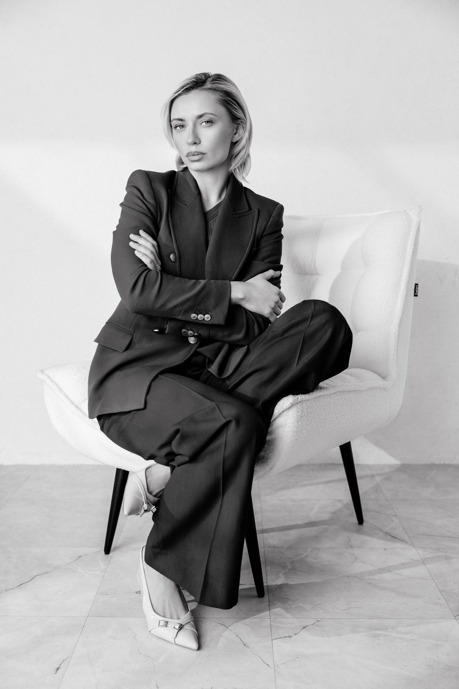 Black and white photo of a confident woman in a tailored suit sitting cross-armed in a modern white chair.