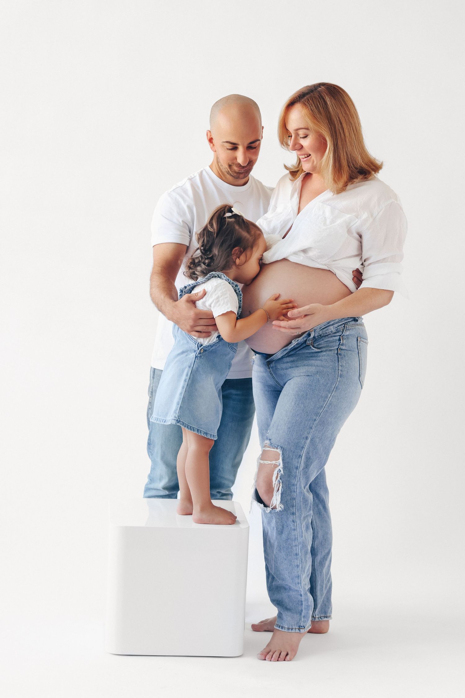 Happy family with bald father, pregnant mother, and young daughter posing together in white and denim outfits on white background.