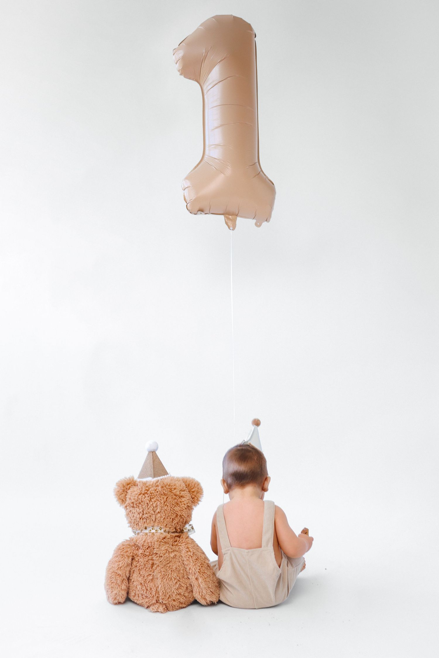 Baby and teddy bear with party hats sit facing a large number 1 balloon on white background.