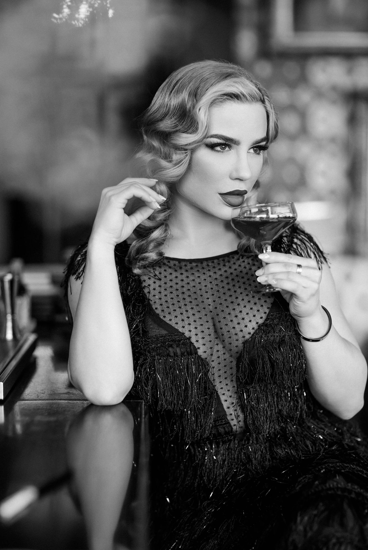 Black and white photo of elegant blonde woman in fringed dress holding cocktail glass at a bar.