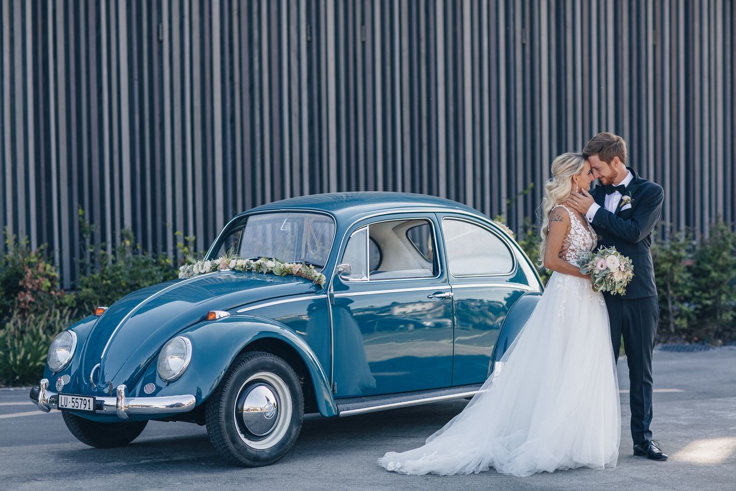 Bride and groom kissing beside a vintage blue VW Beetle decorated with flowers at their wedding.