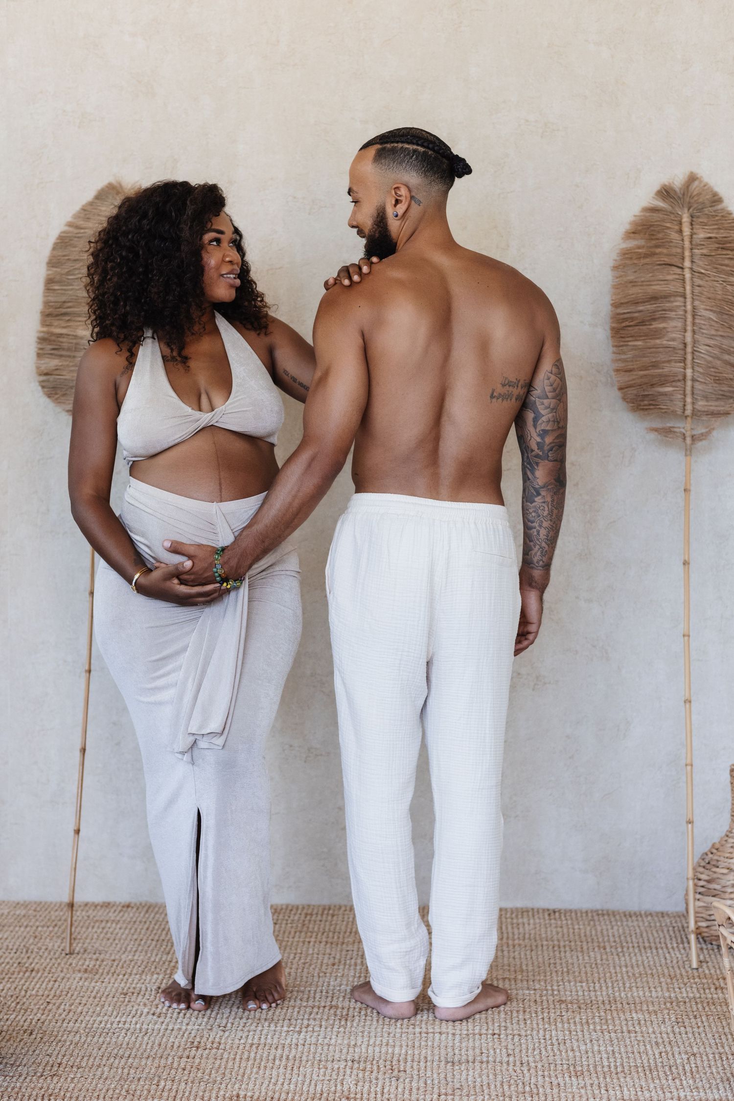 Pregnant Black woman in white crop top and skirt poses with shirtless tattooed man in white pants indoors.