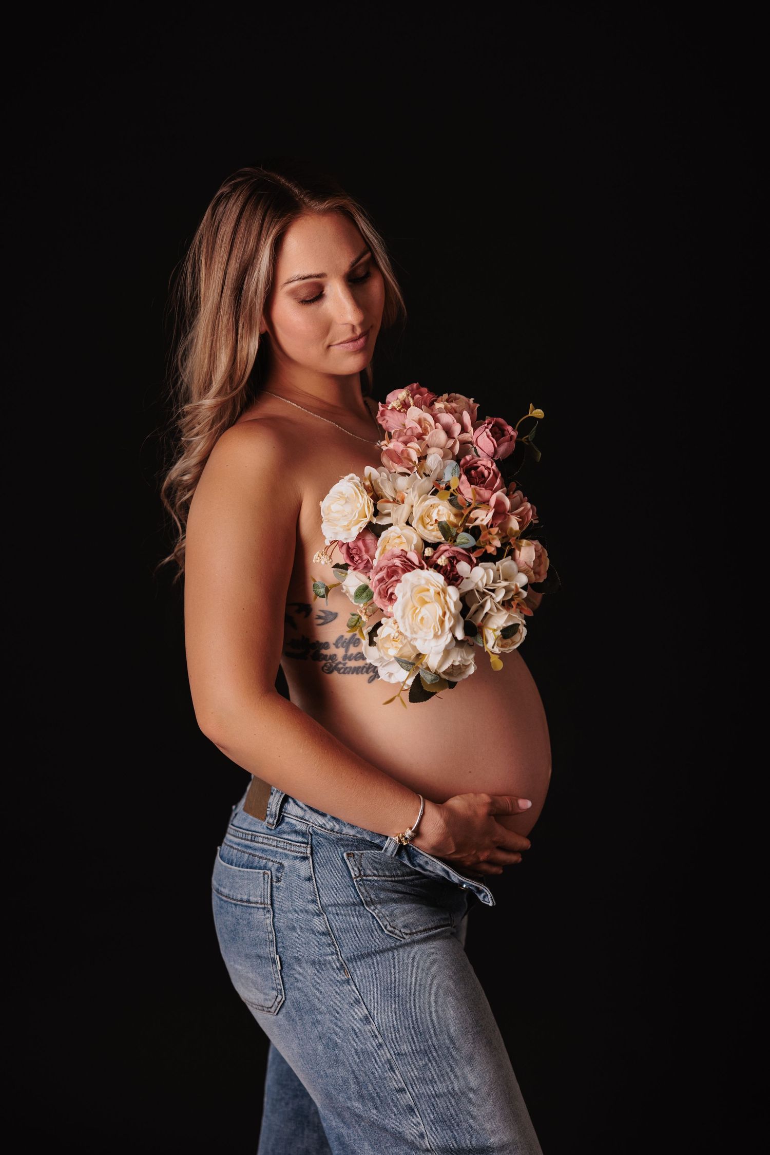 Pregnant woman in jeans holding floral bouquet over chest, posing against black background in maternity photo shoot.