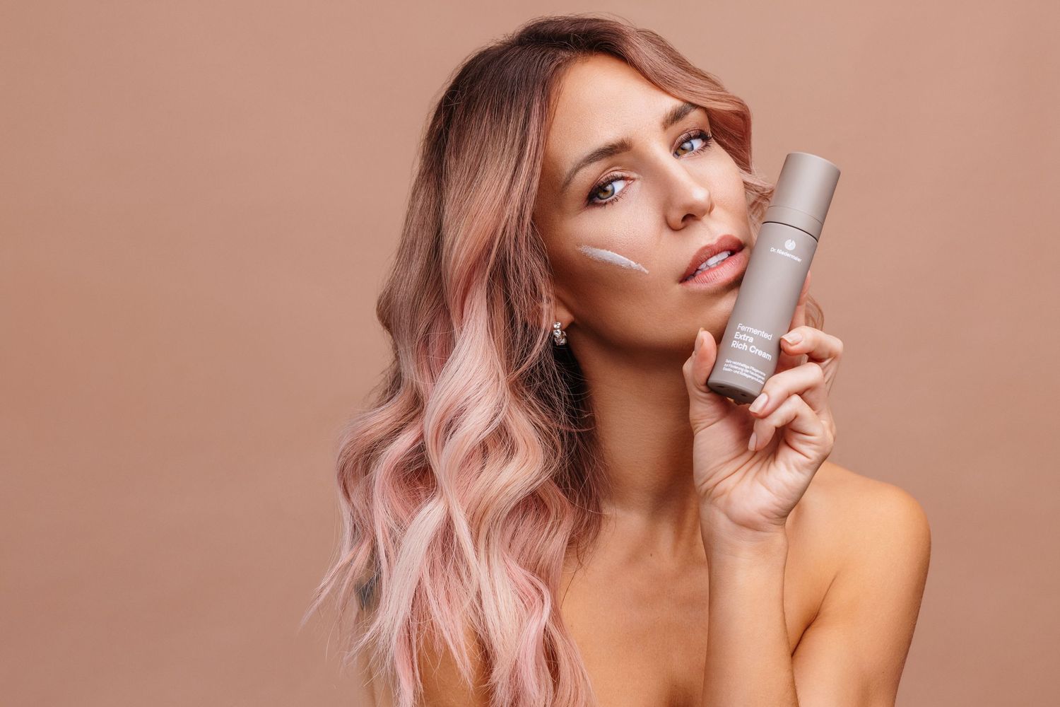 Woman with pink wavy hair holding a skincare product tube, with cream applied on her cheek, against beige background.