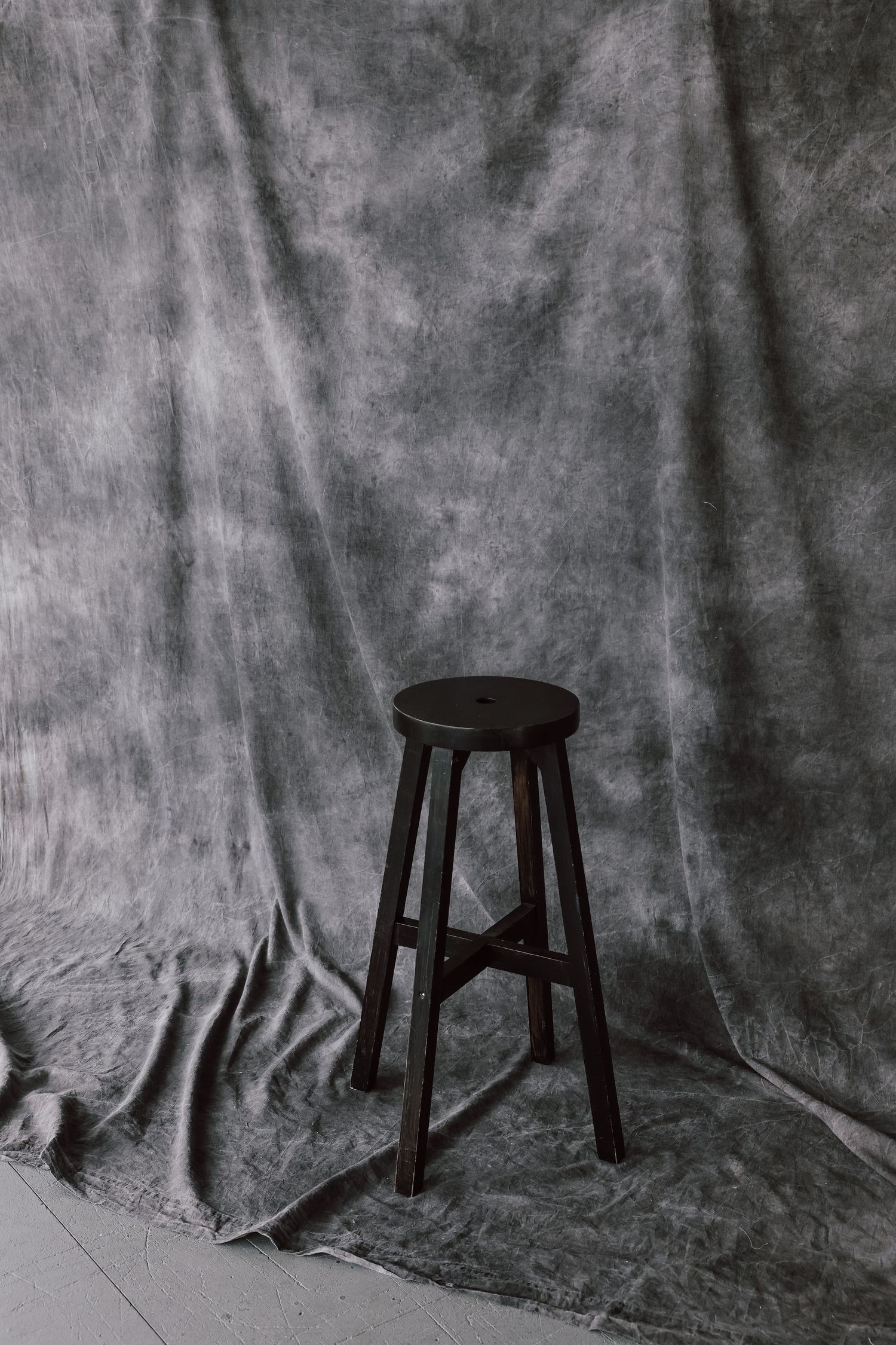 A dark wooden bar stool stands alone against a mottled gray fabric backdrop in a photography studio.