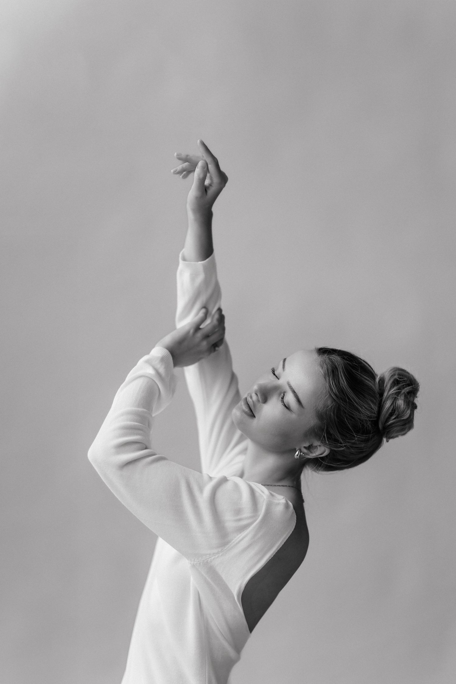 Black and white photo of a graceful dancer in white outfit with arms raised and eyes closed against light background.