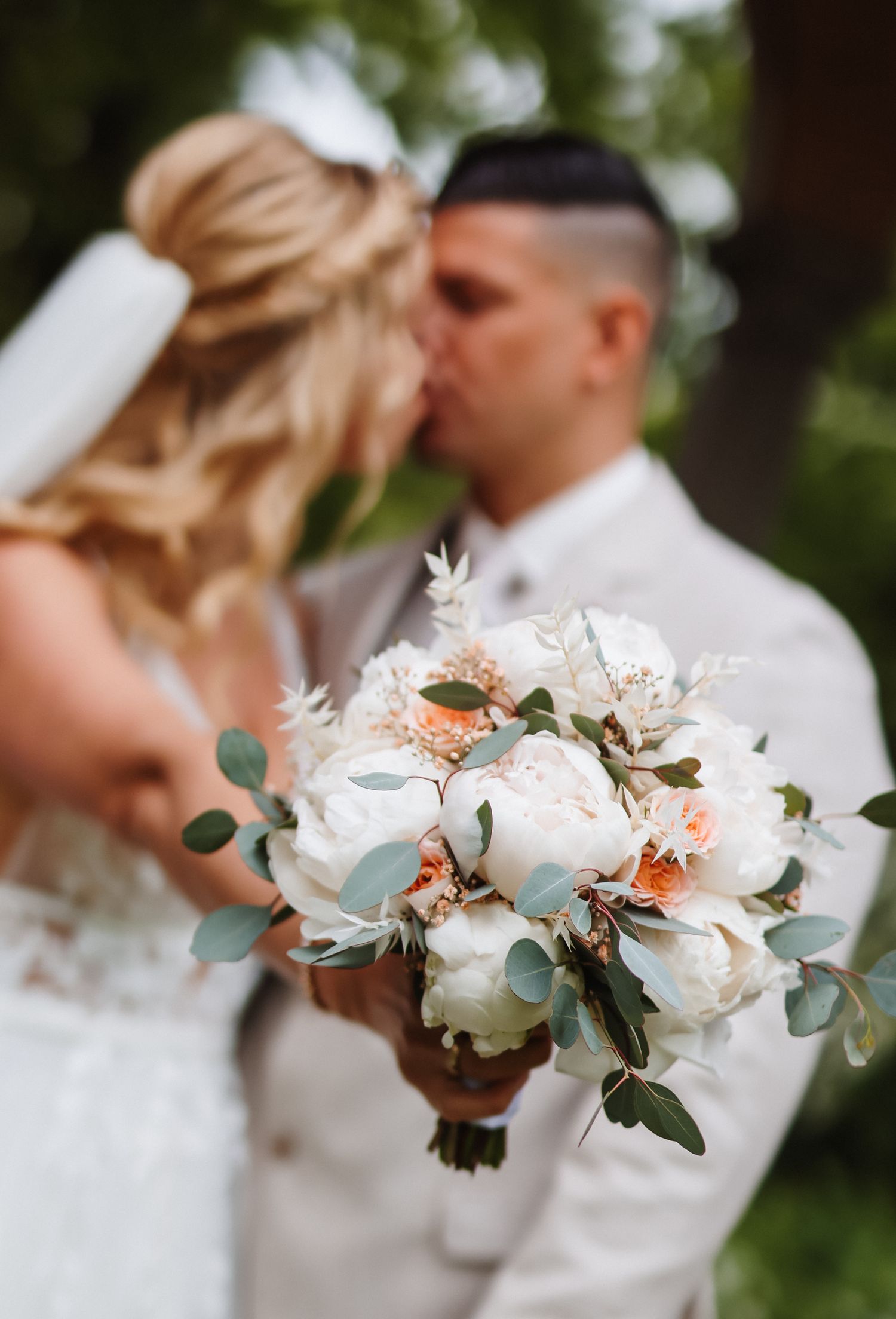 Bride and groom kissing behind a white peony and eucalyptus wedding bouquet with soft peach accents outdoors.