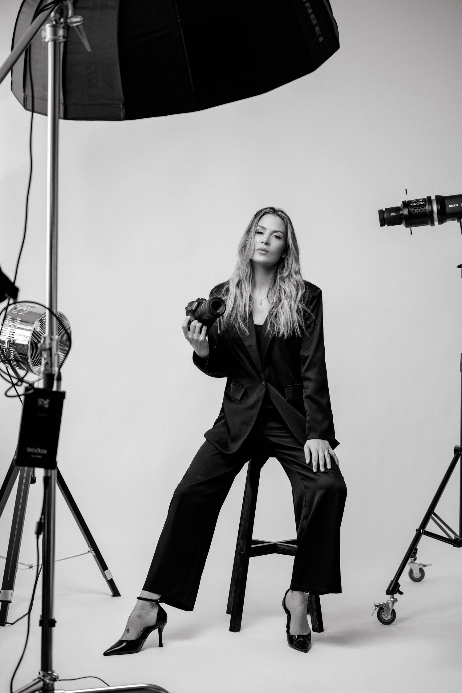 Black and white photo of a blonde woman in a black suit holding a camera, seated on a stool in a photography studio.