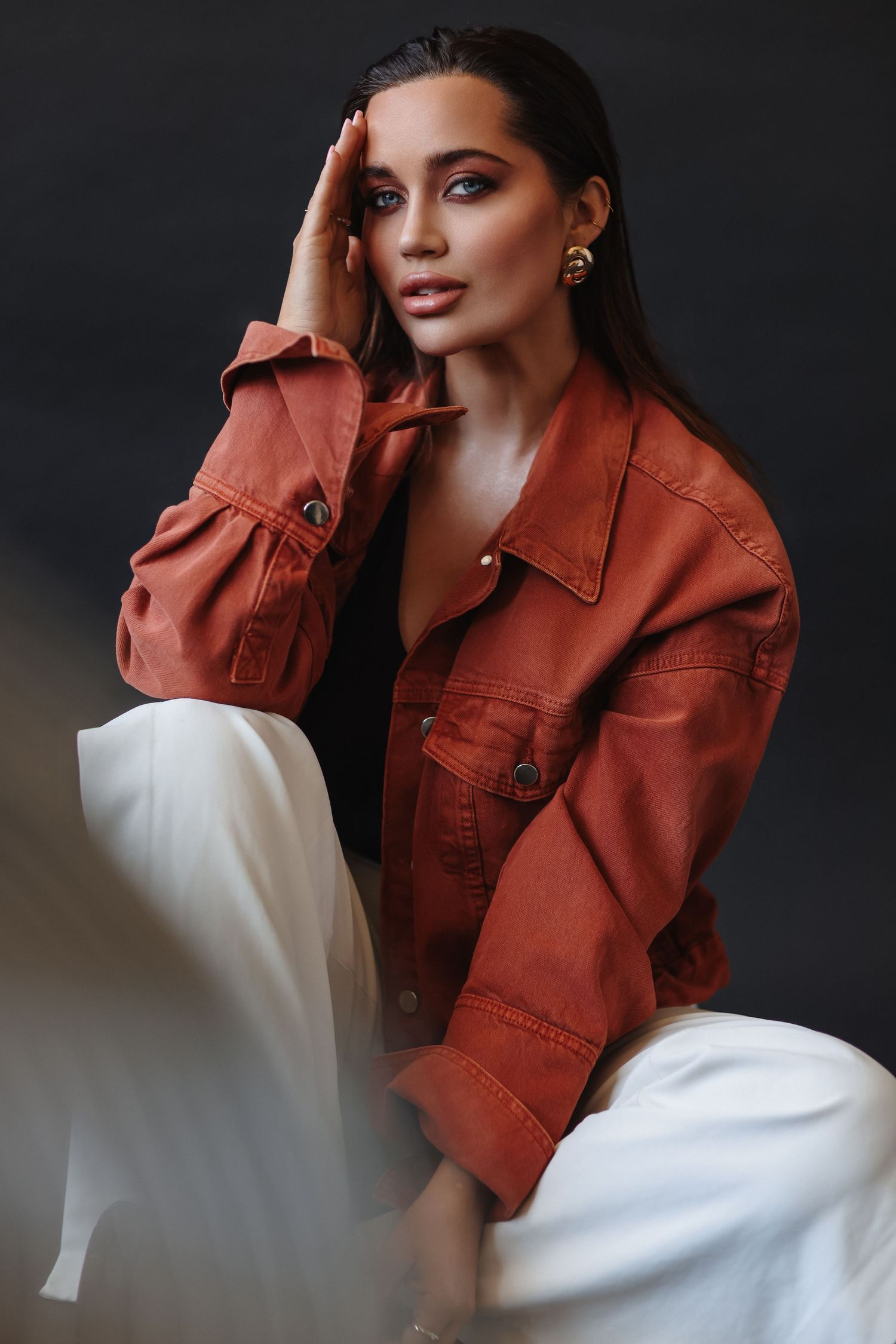 Woman in rust orange denim jacket and white wide-leg pants poses seated against dark studio background.