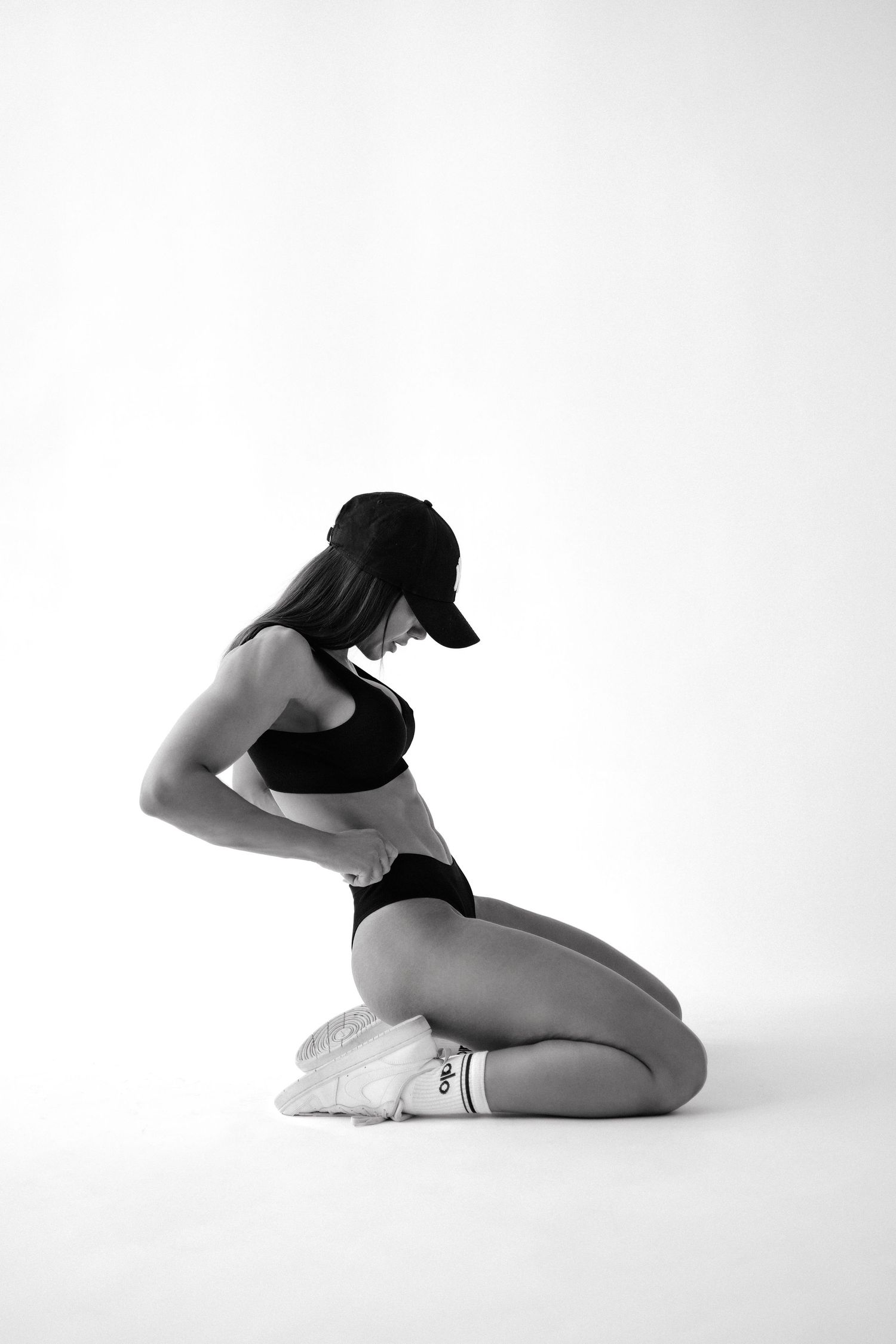 Black and white photo of a fit woman in athletic wear kneeling on the floor, wearing a cap and sneakers.