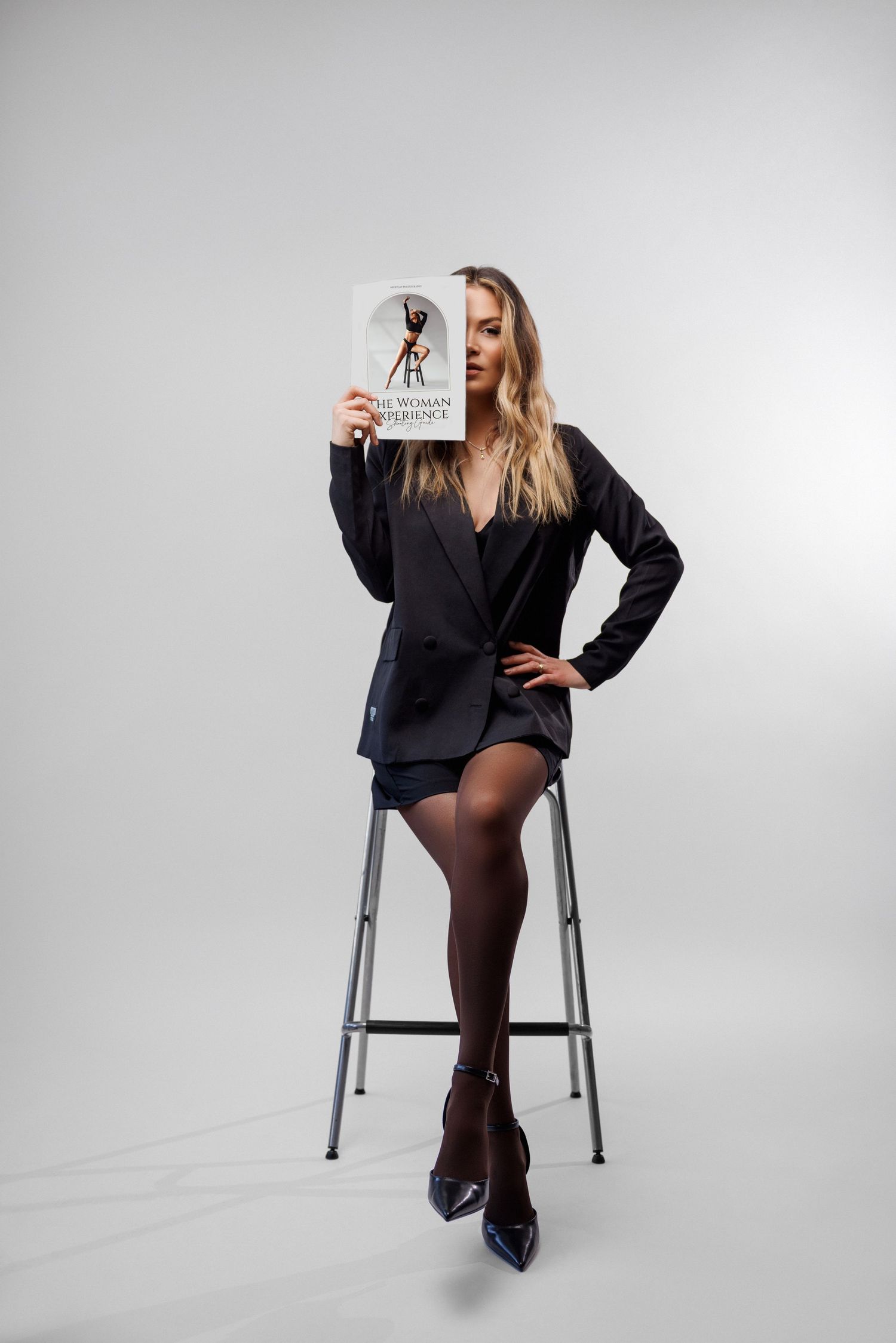 Woman in black blazer and tights holding magazine on stool against white background.