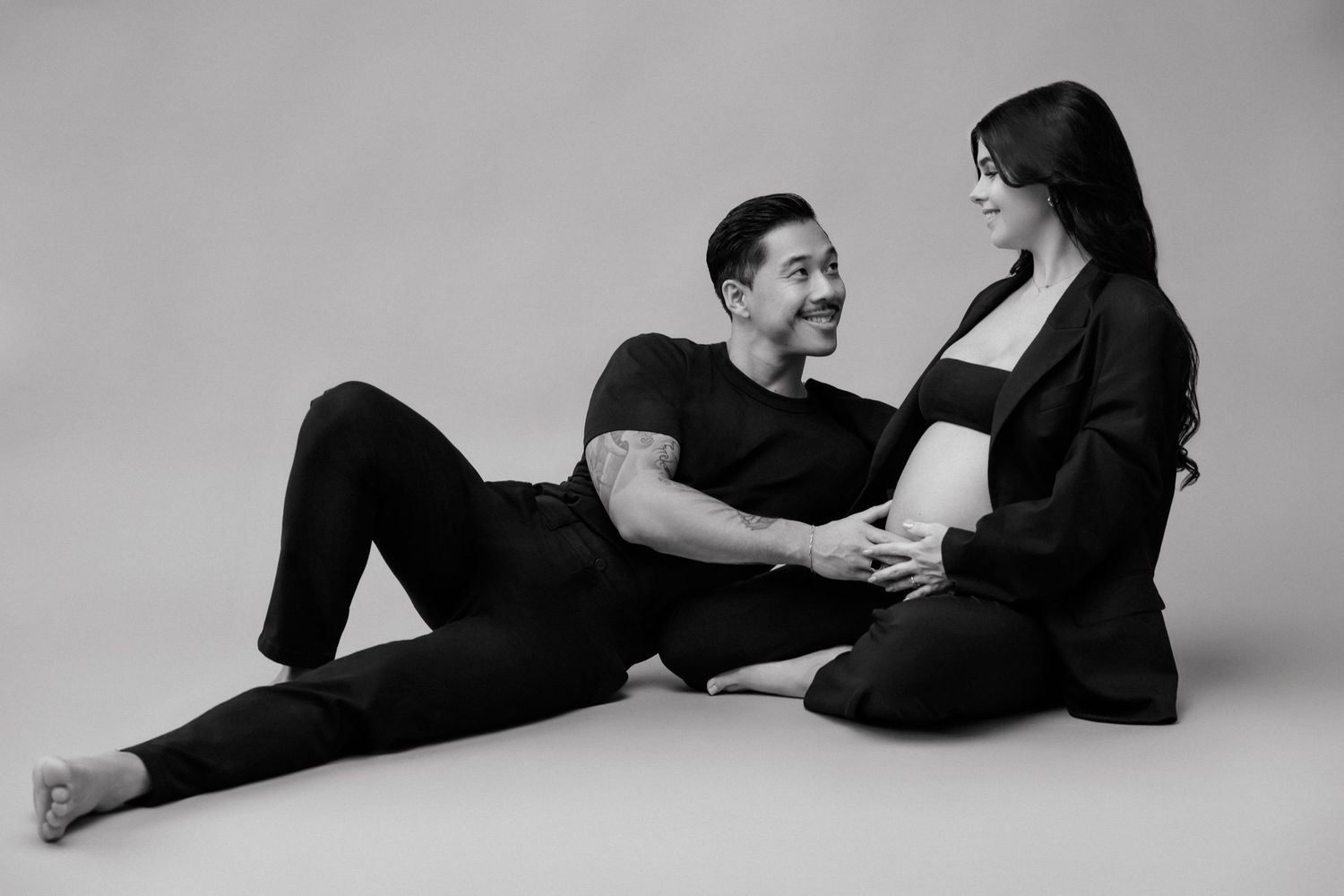 Black and white maternity photo of a couple sitting on floor, man smiling at pregnant woman cradling her baby bump.