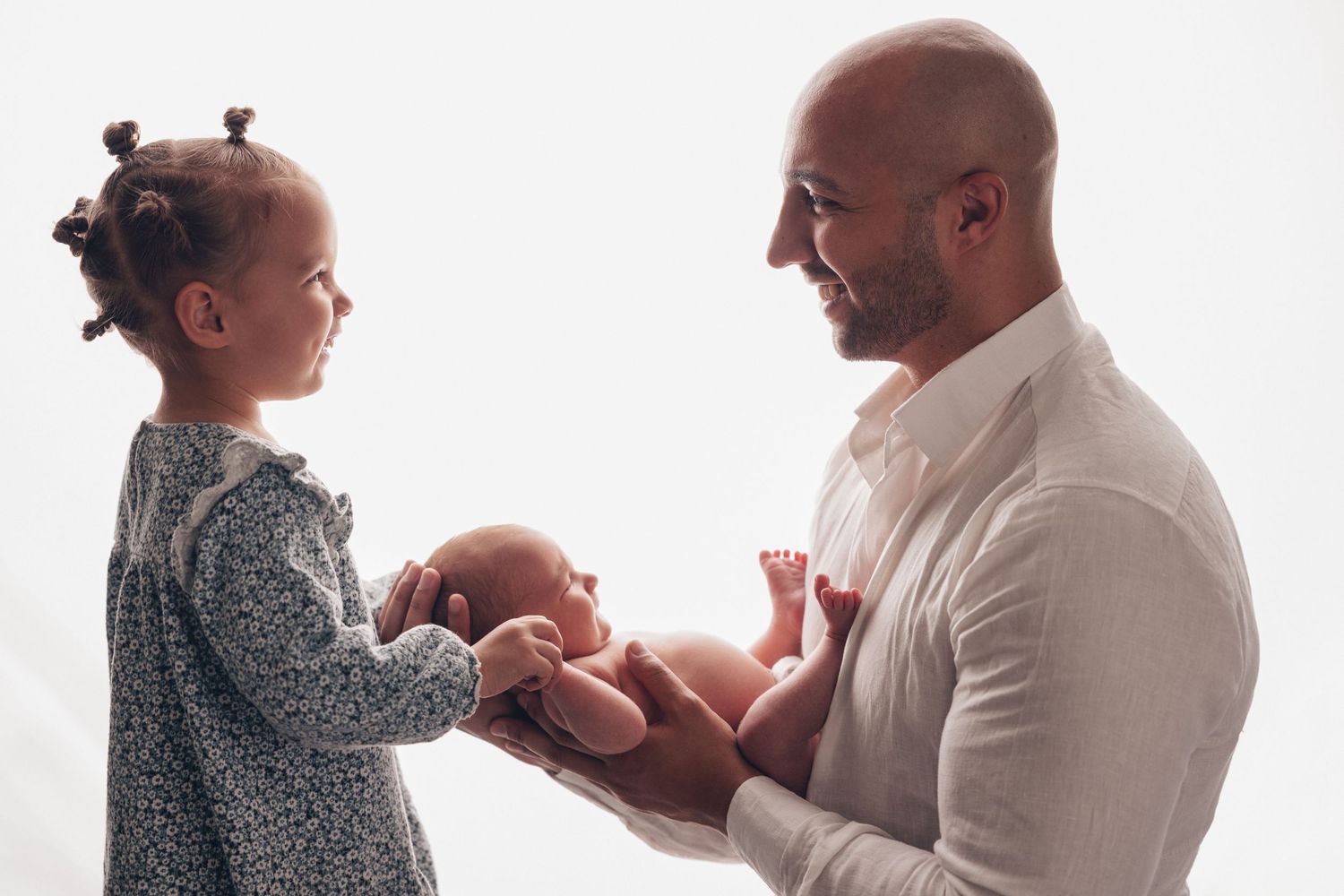 Bald father holds newborn baby while young toddler girl in gray dress looks on, smiling, against white background.