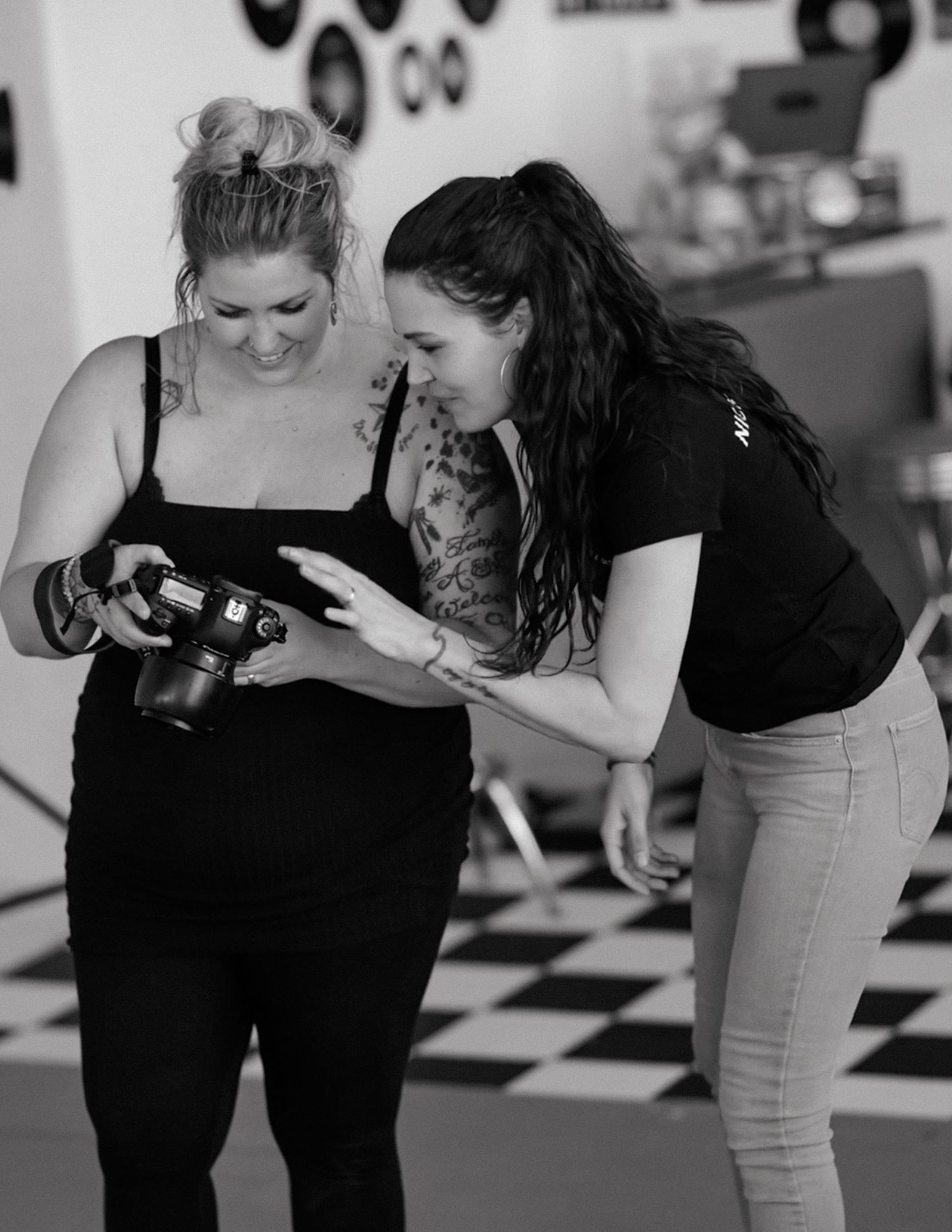 Two women in black outfits review photos on a DSLR camera in a retro studio with checkered floors.