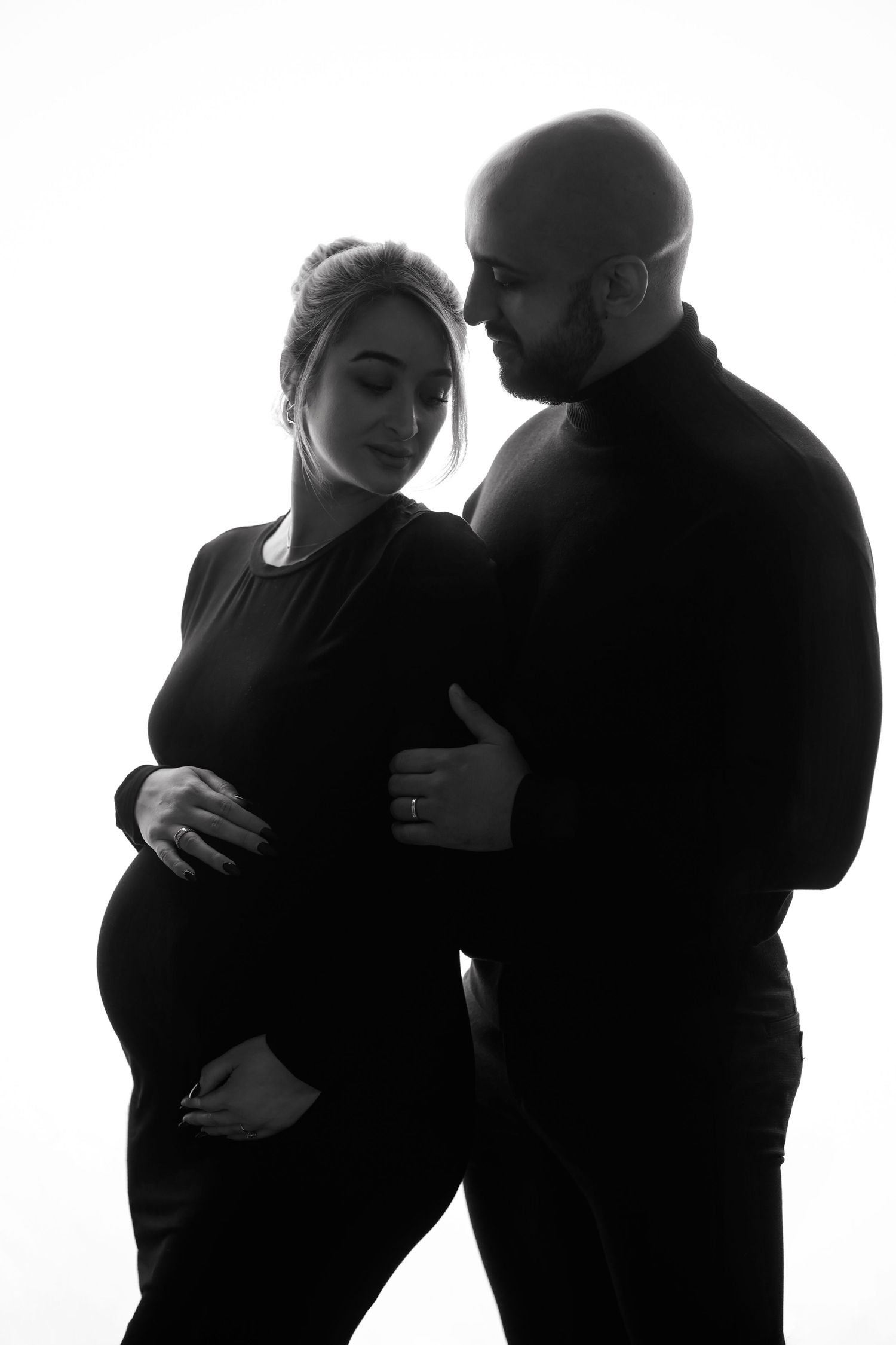 Black and white maternity photo of a couple, man embracing pregnant woman from behind in a tender moment.