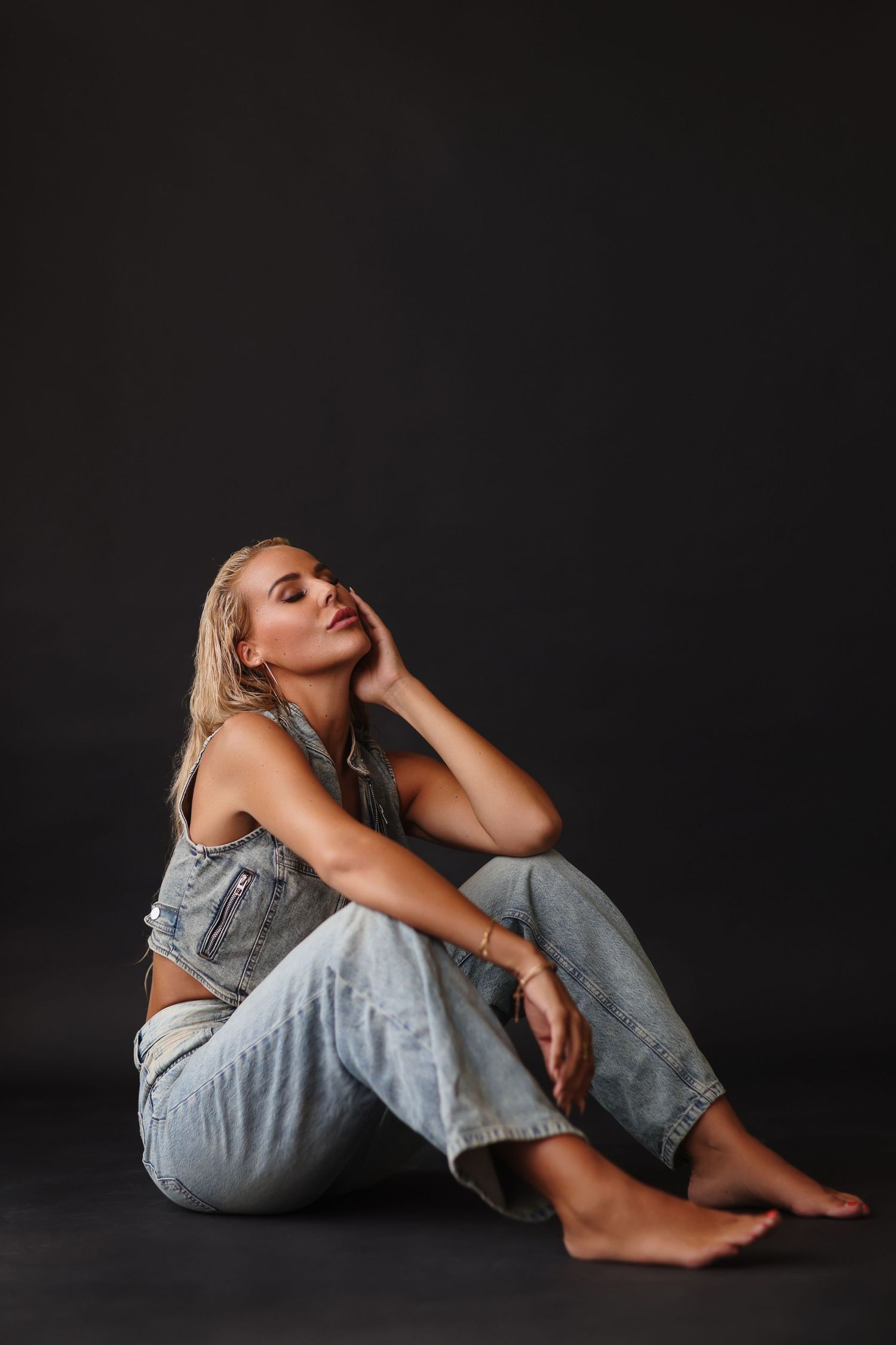 Blonde woman sitting barefoot on dark floor in denim outfit, posing thoughtfully with hand on face.