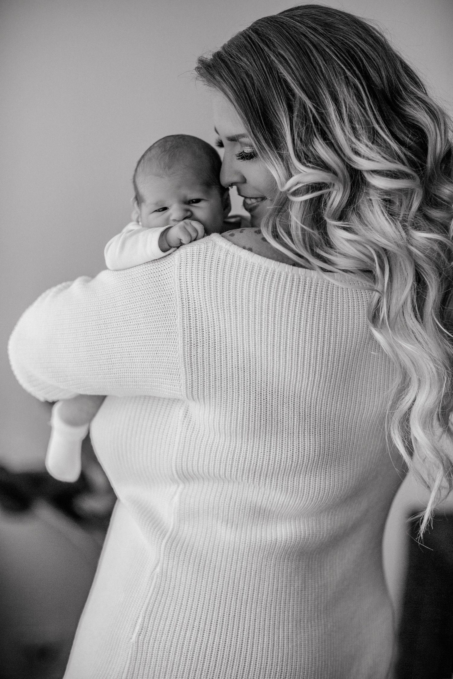 Black and white photo of a mother with wavy blonde hair holding her newborn baby close in a white sweater.