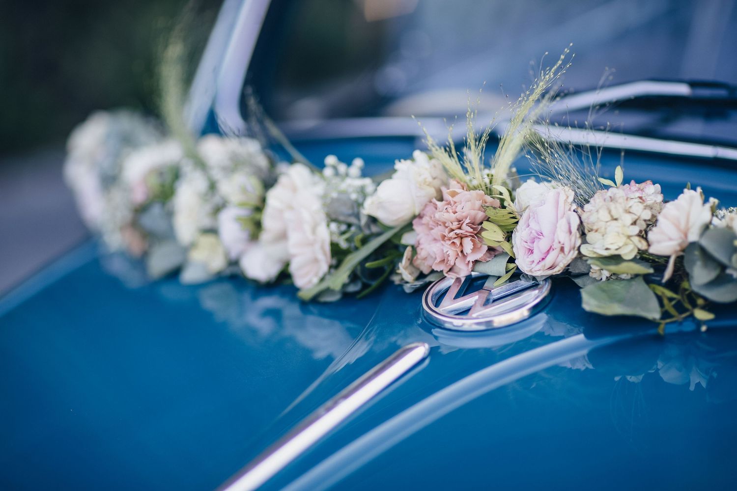 Blue vintage wedding car hood adorned with elegant floral arrangement of roses, carnations, and greenery with decorative rings.