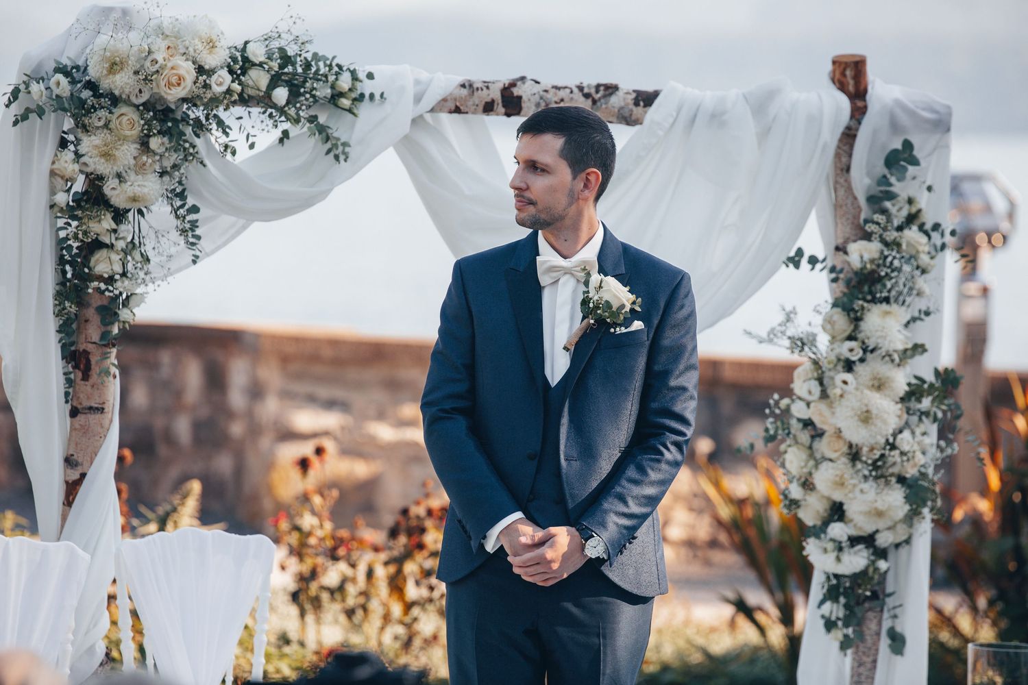 Groom in navy suit standing at outdoor wedding altar decorated with white flowers and draped fabric.