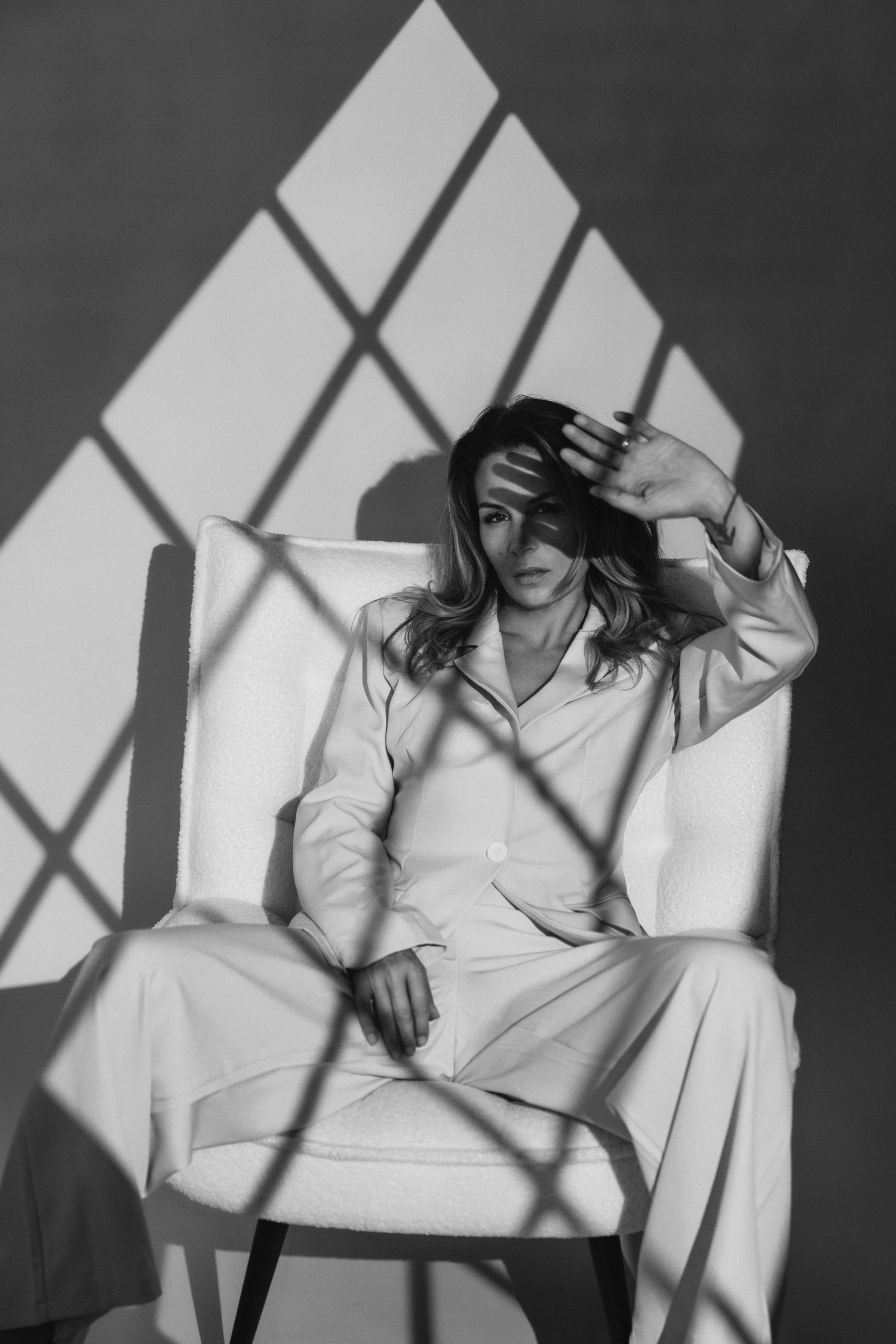 Black and white portrait of a woman in light pajamas sitting on an armchair, raising her hand to shield her face from window shadows.