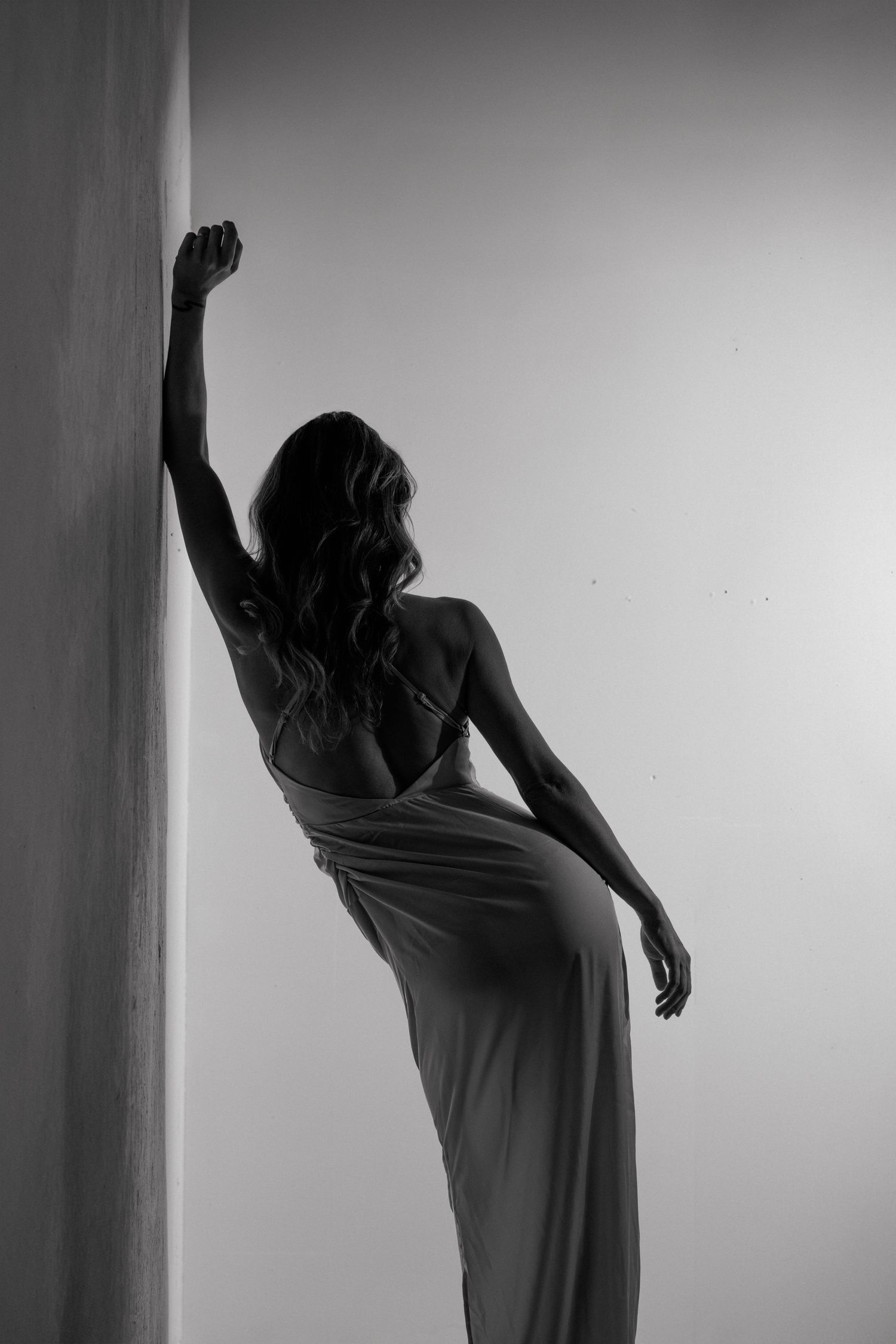 Black and white silhouette of a woman in a flowing gown leaning against a wall with one arm raised, viewed from behind.