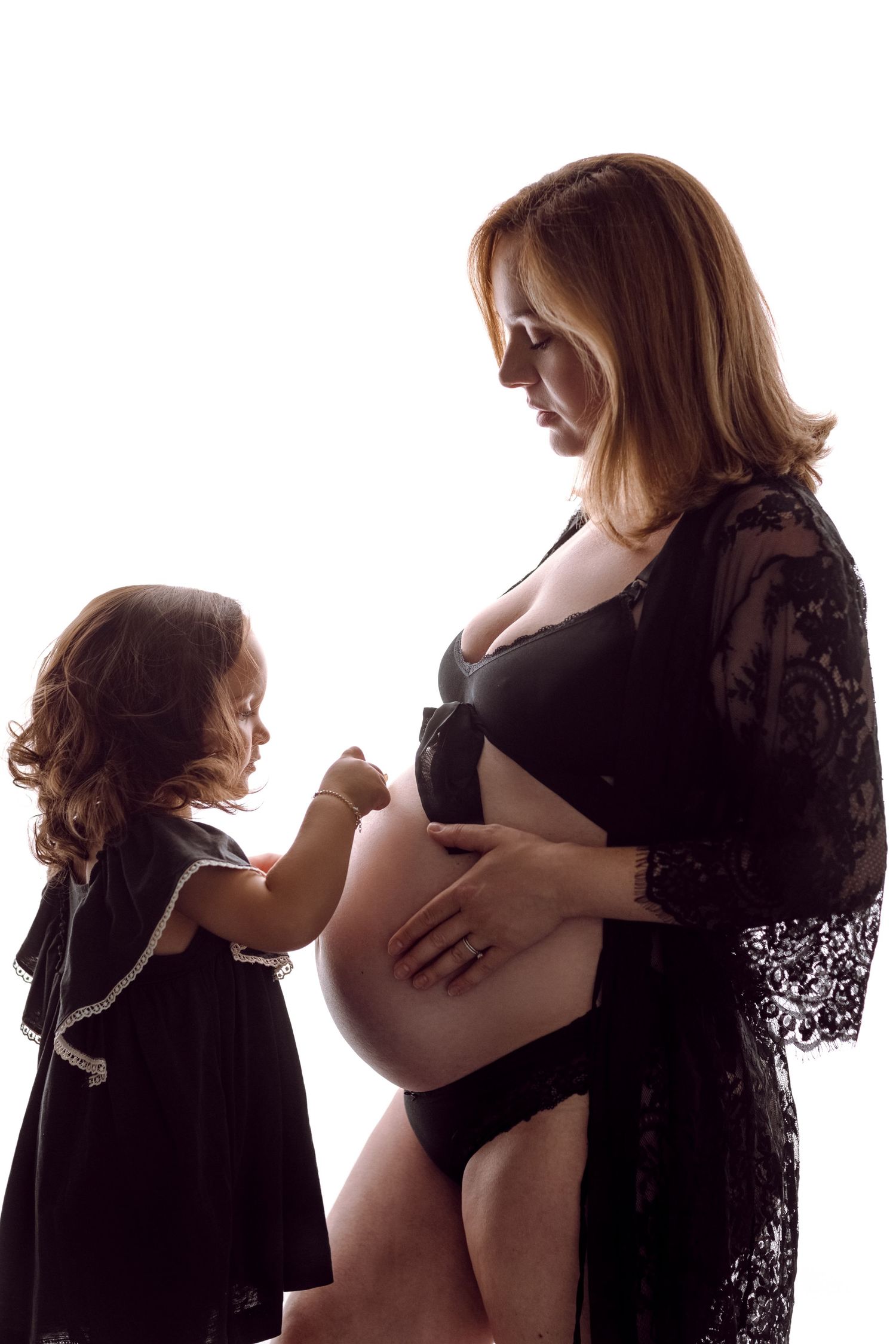 Pregnant woman in black lace robe with toddler girl touching her baby bump in a tender maternity photo.