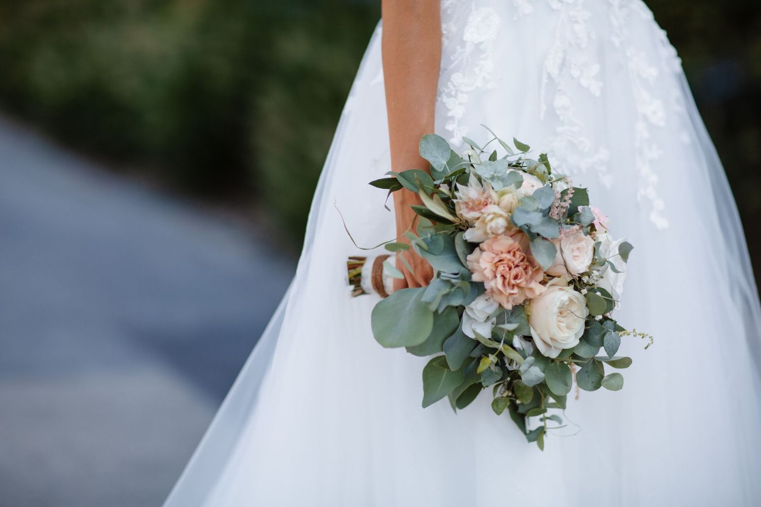 Bride in white lace gown holding elegant bouquet of blush pink, ivory roses and eucalyptus greenery.