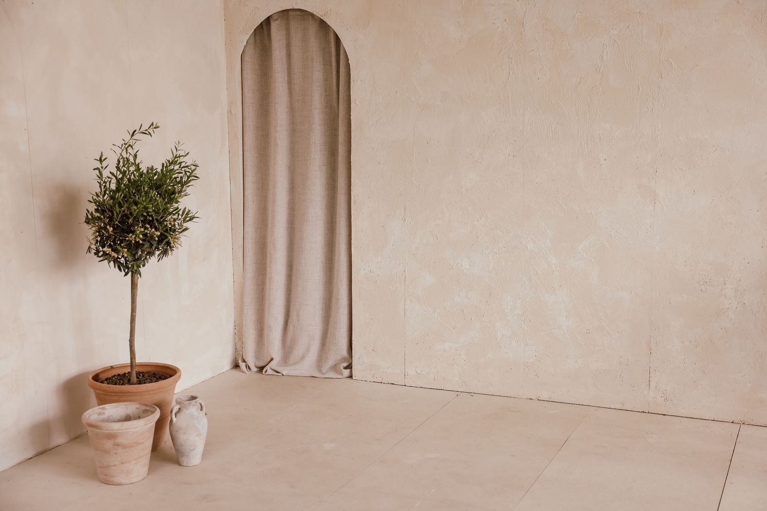 Minimalist beige room with arched linen curtain, terracotta potted olive tree, and small ceramic vase on tiled floor.