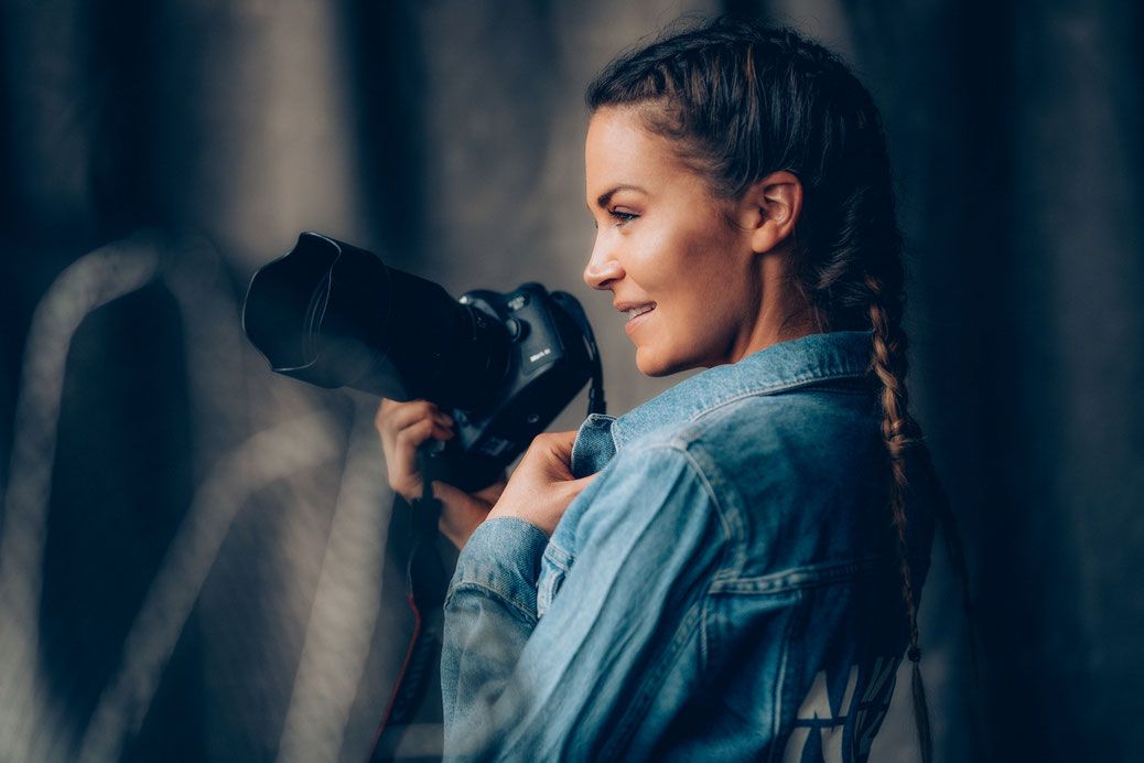 Young woman photographer in denim jacket smiling while holding a professional DSLR camera outdoors.