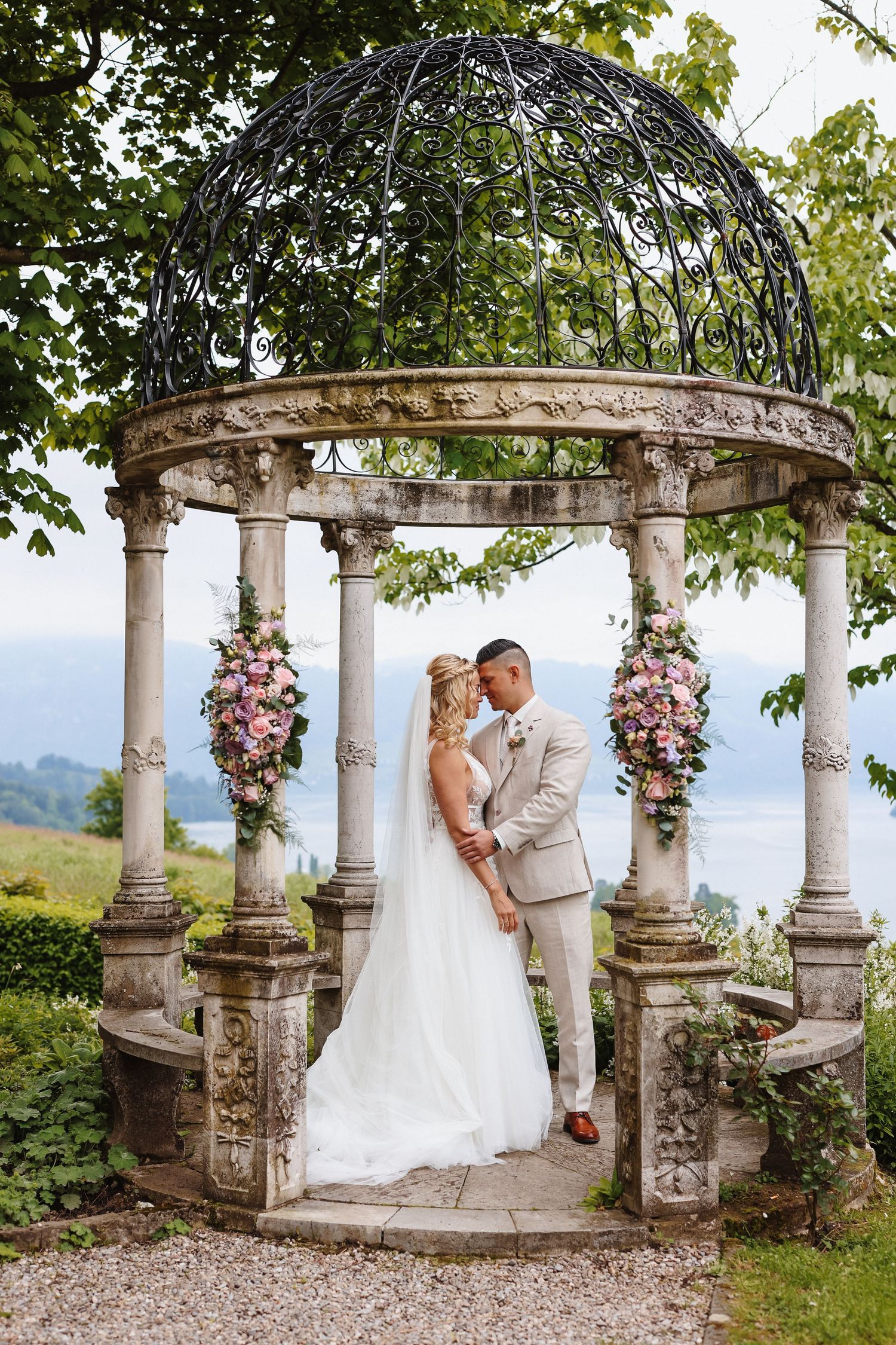 Bride and groom share a romantic kiss under an ornate stone gazebo adorned with pink flowers, with scenic views behind them.