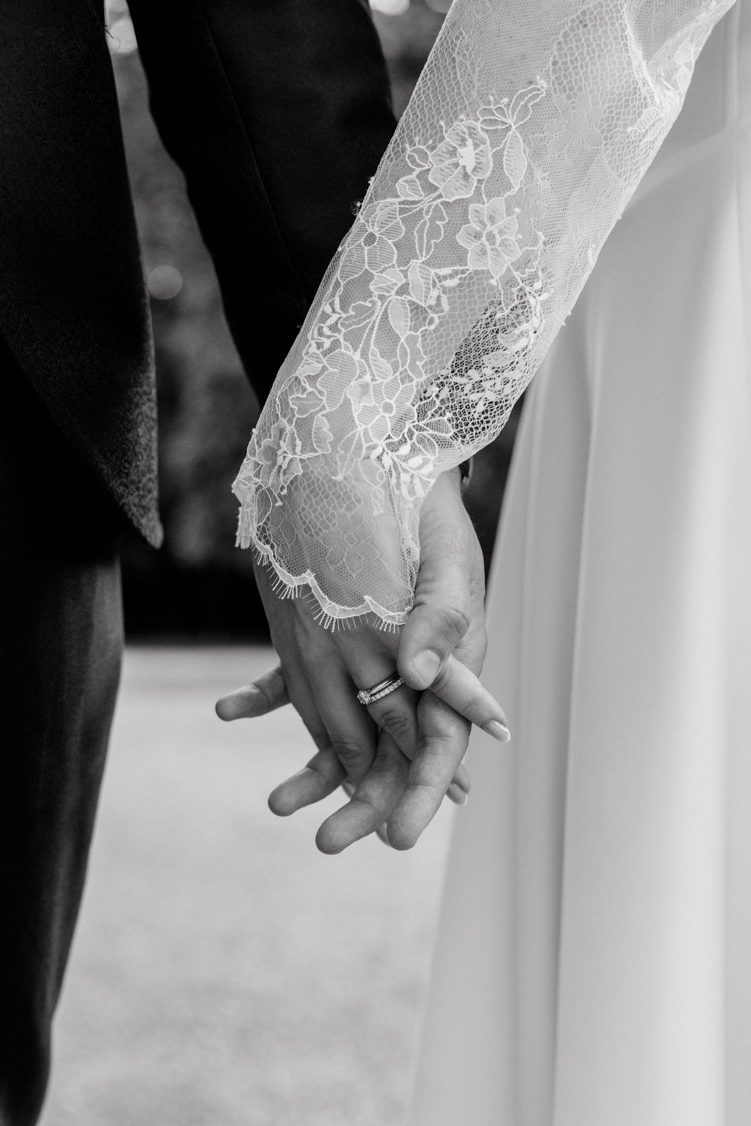 Black and white photo of bride and groom holding hands, showcasing wedding rings and lace bridal sleeve.