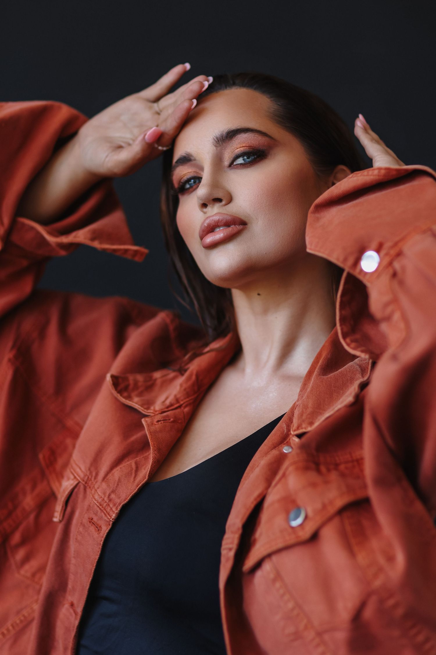 Woman in rust orange denim jacket over black bodysuit posing confidently against dark background.