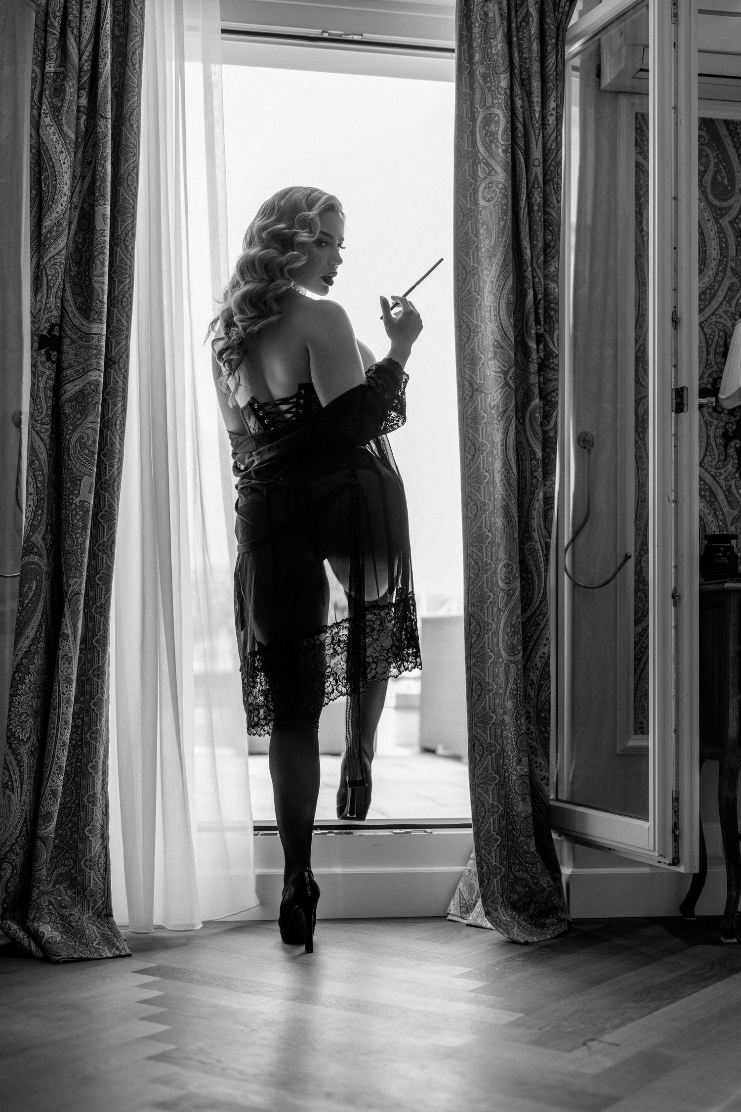 Black and white photo of elegant woman in lingerie holding cigarette holder, standing by open window with curtains.